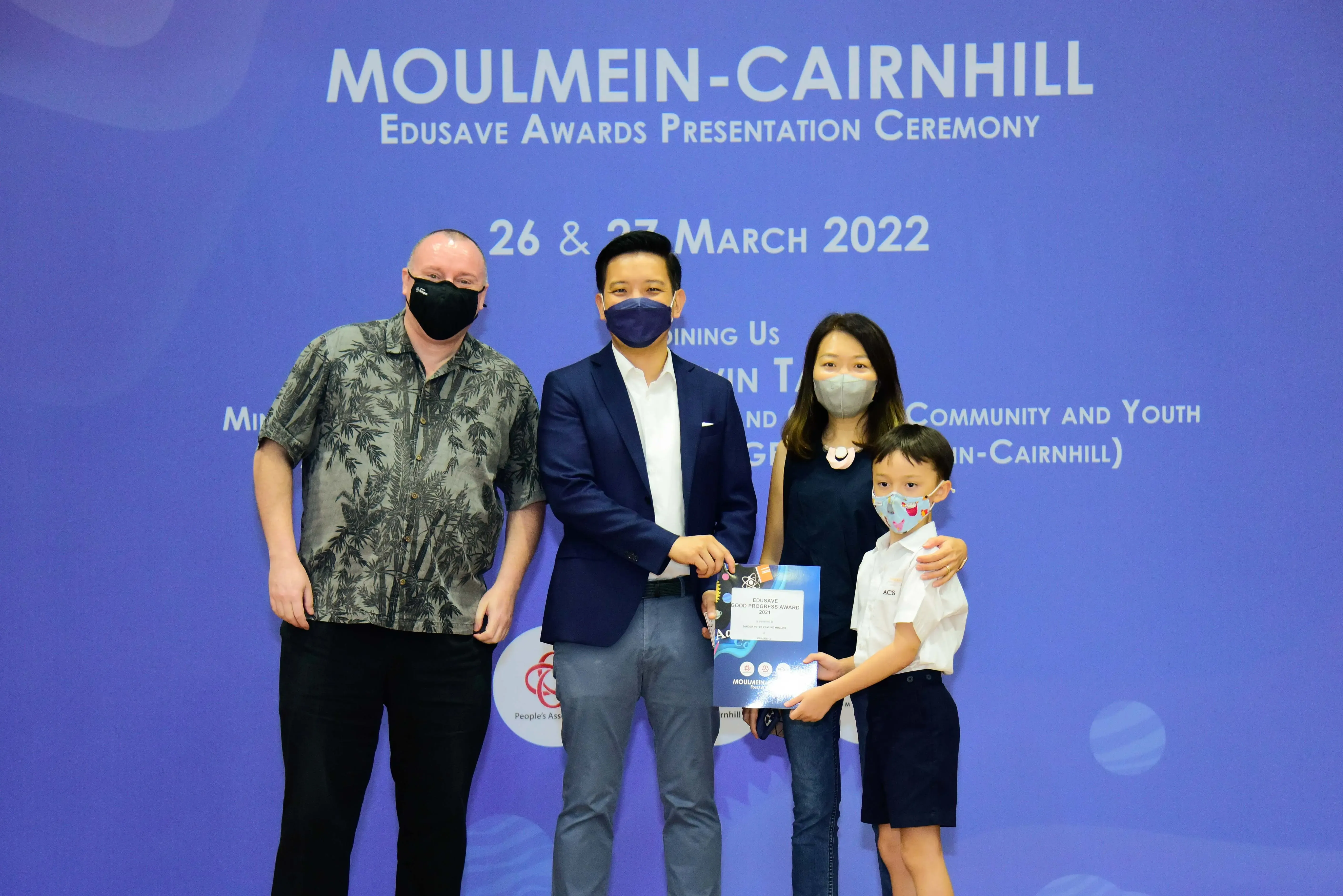 Four masked people pose at the Moulmein-Cairnhill Edusave Awards Ceremony on 26-27 March 2022, with a child holding a certificate.