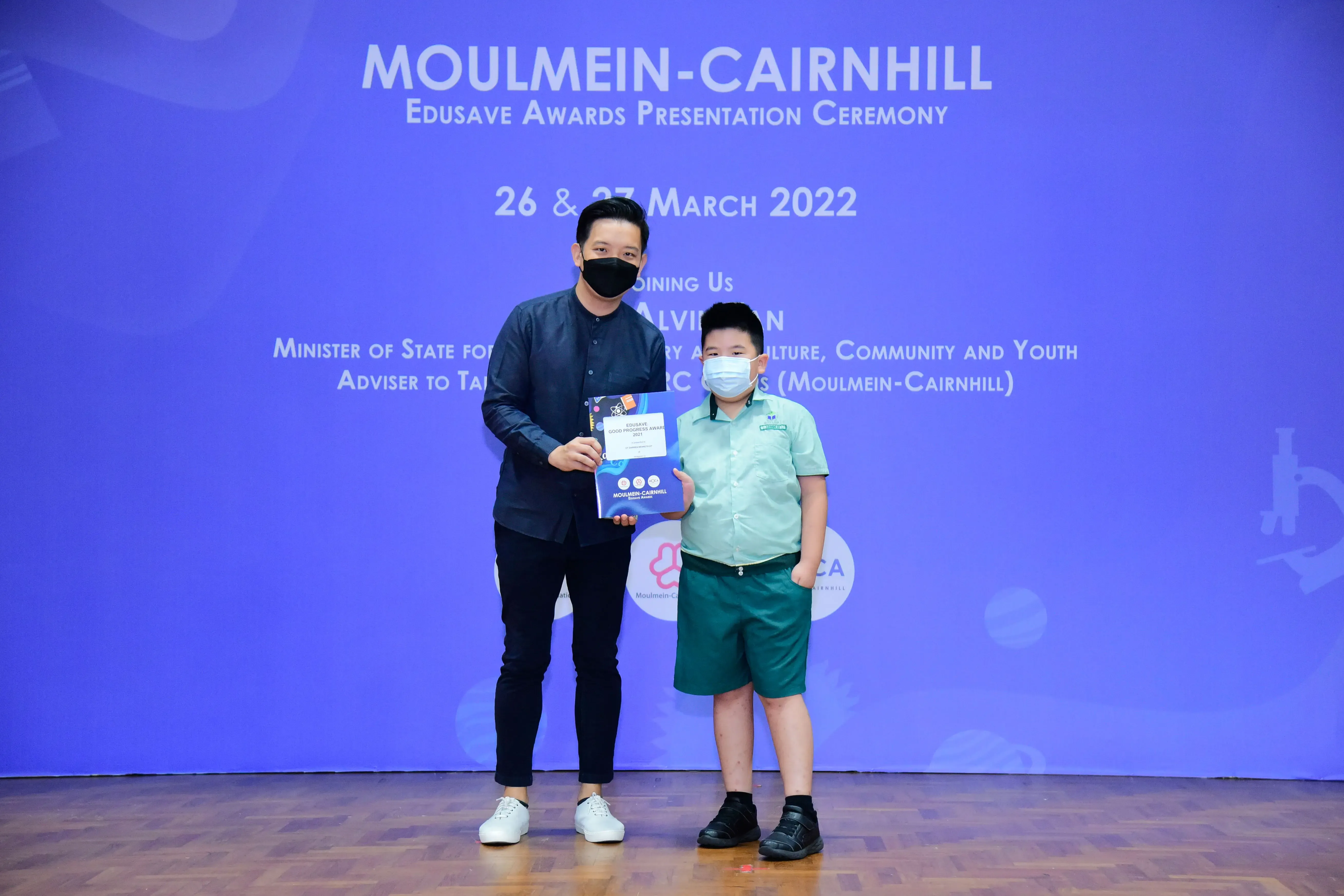 An adult and a young boy in school uniform hold an award certificate at the Moulmein-Cairnhill Edusave Awards Ceremony 2022.