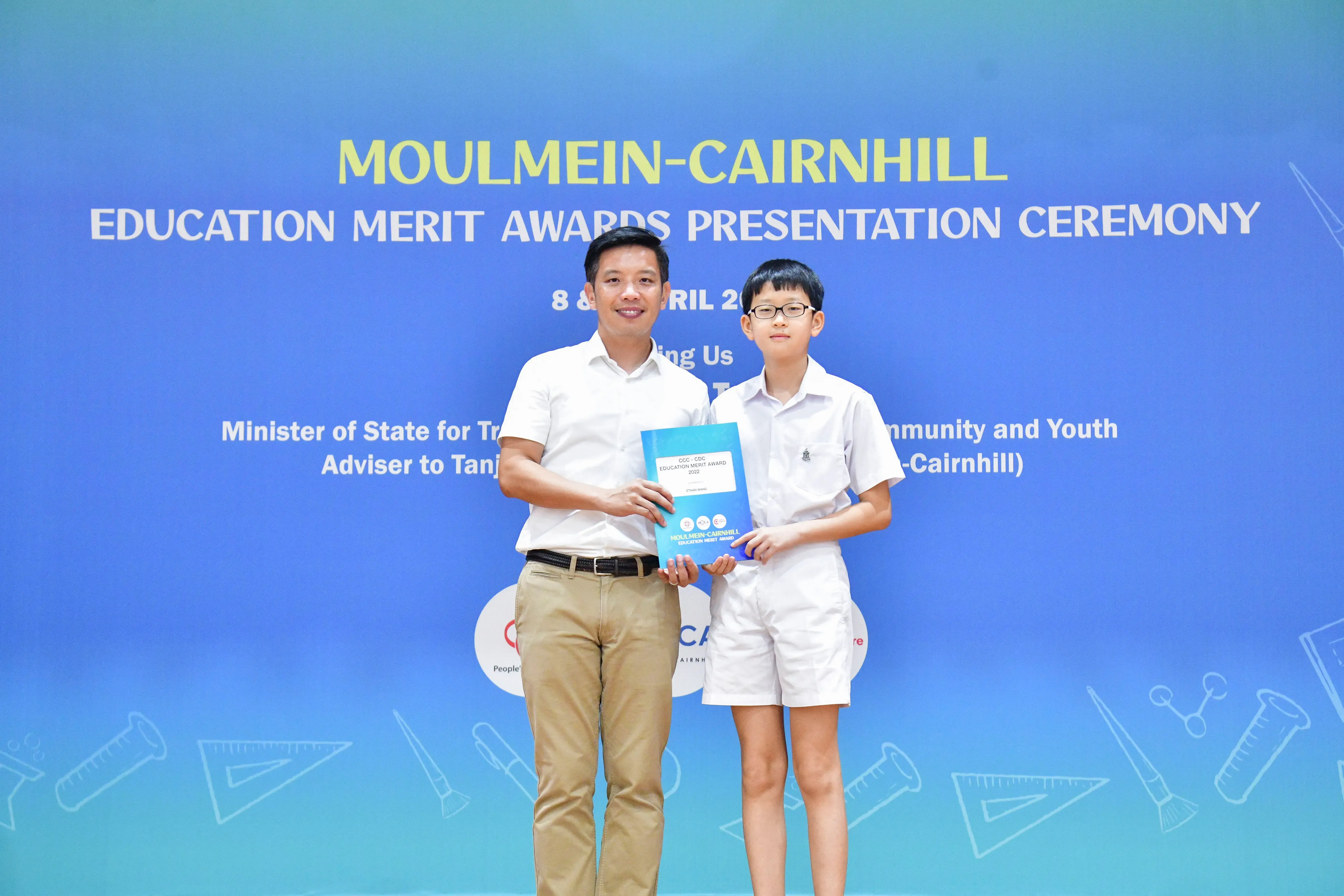 An adult man and a young boy hold a blue award booklet at the Moulmein-Cairnhill Education Merit Awards ceremony.