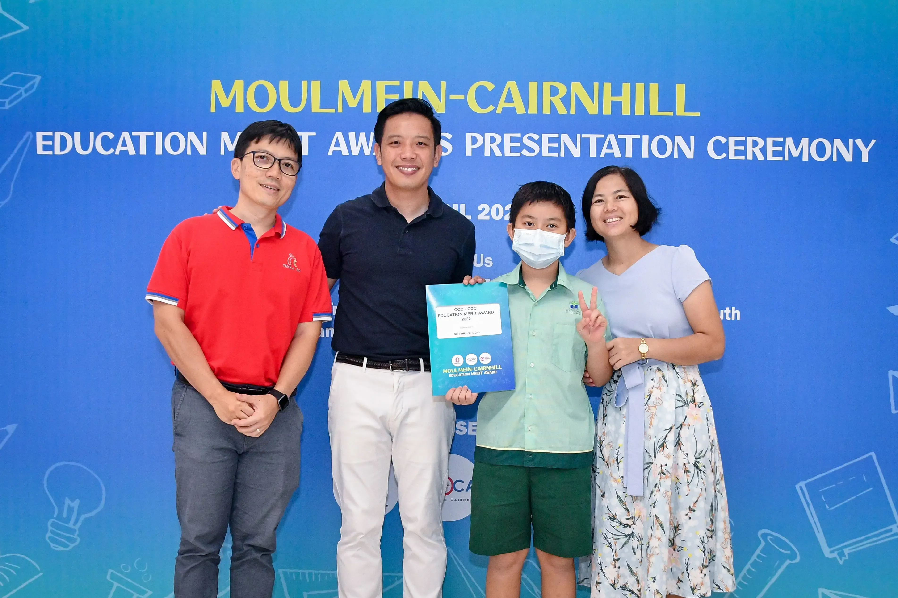 Four people pose before a Moulmein-Cairnhill Education Merit Awards banner; a masked boy holds a certificate.