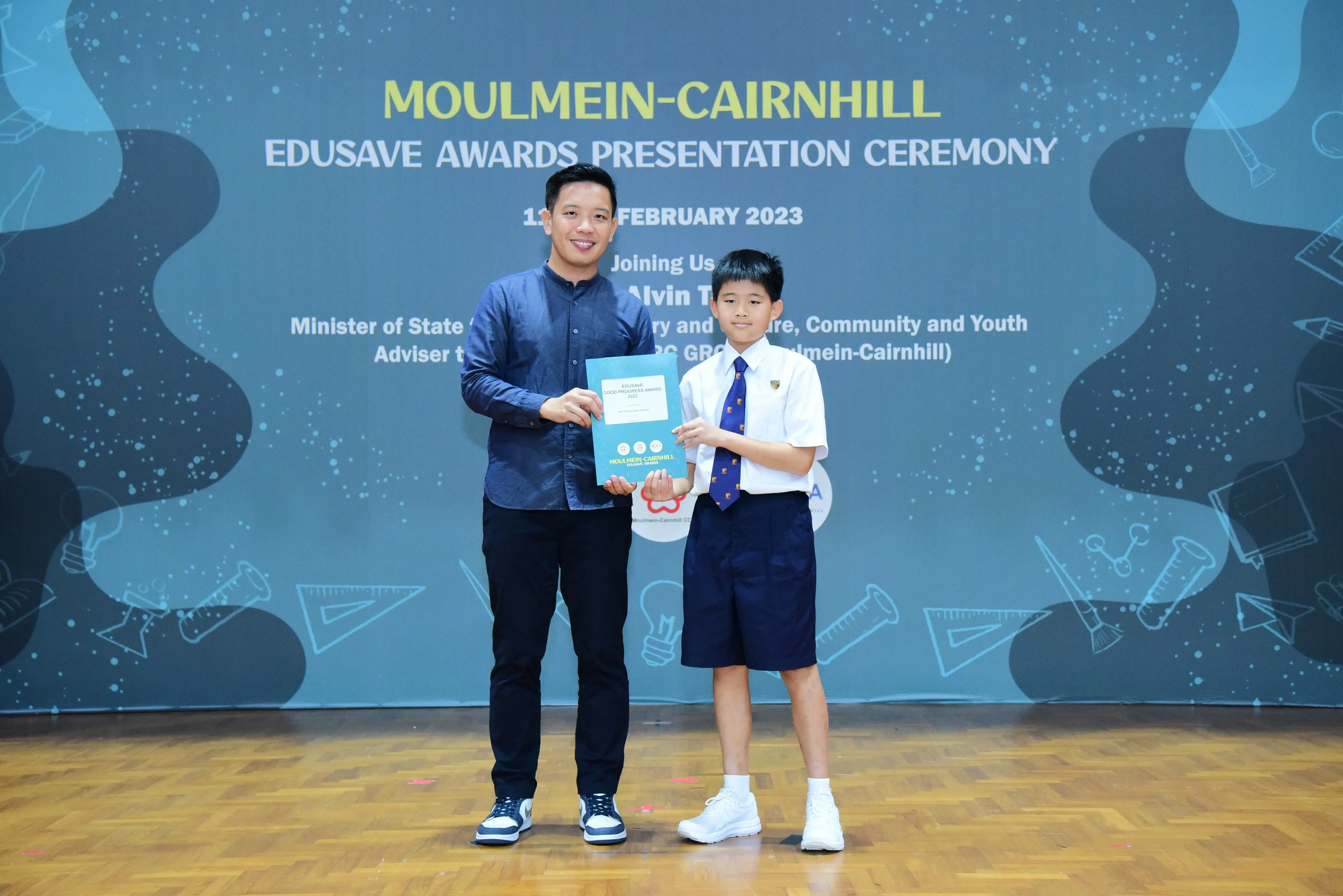 An adult and a young student in uniform hold a certificate at the Moulmein-Cairnhill Edusave Awards Ceremony 2023.