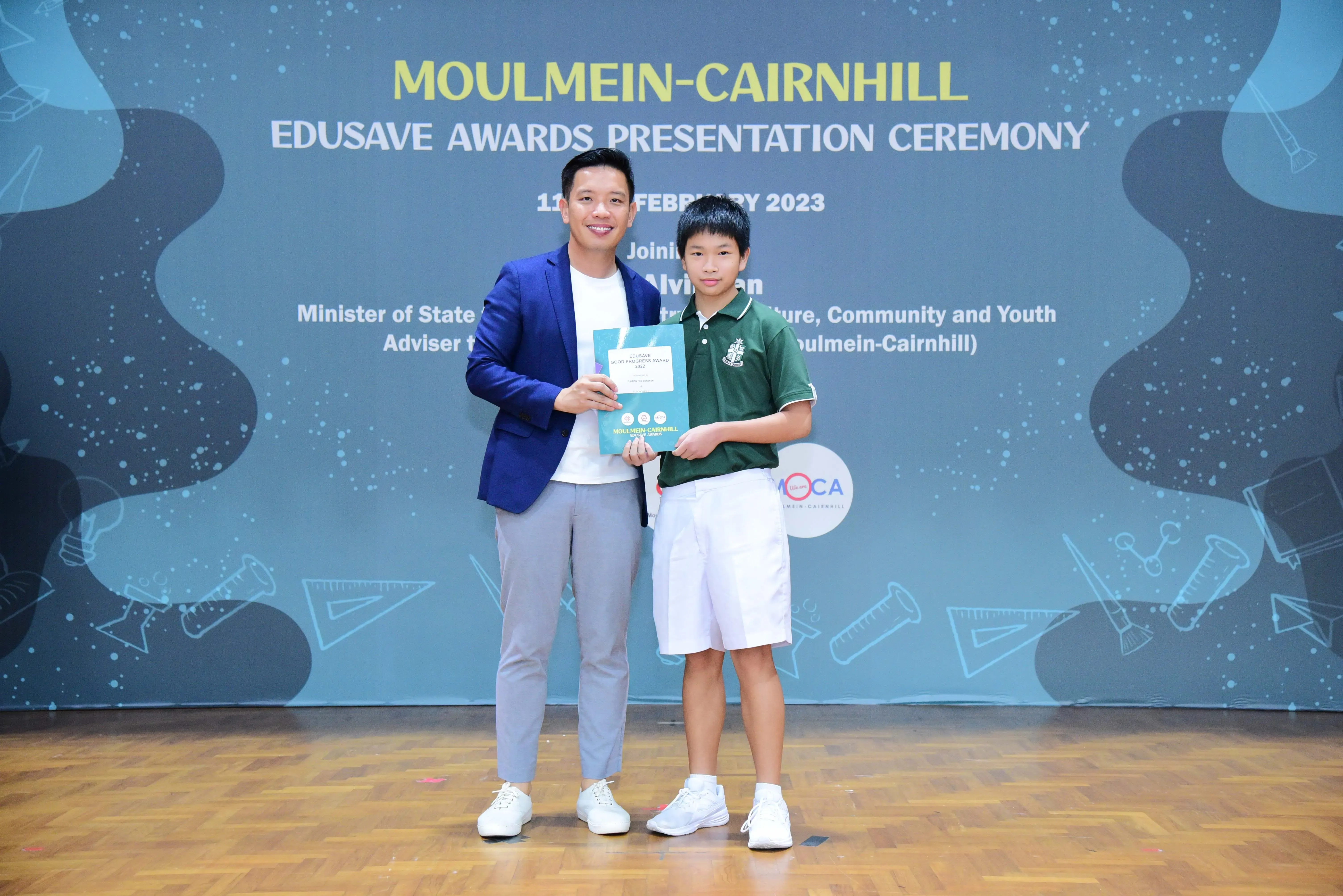 A man in a blue blazer and a young boy in a school uniform hold a certificate at the Moulmein-Cairnhill Edusave Awards Ceremony.