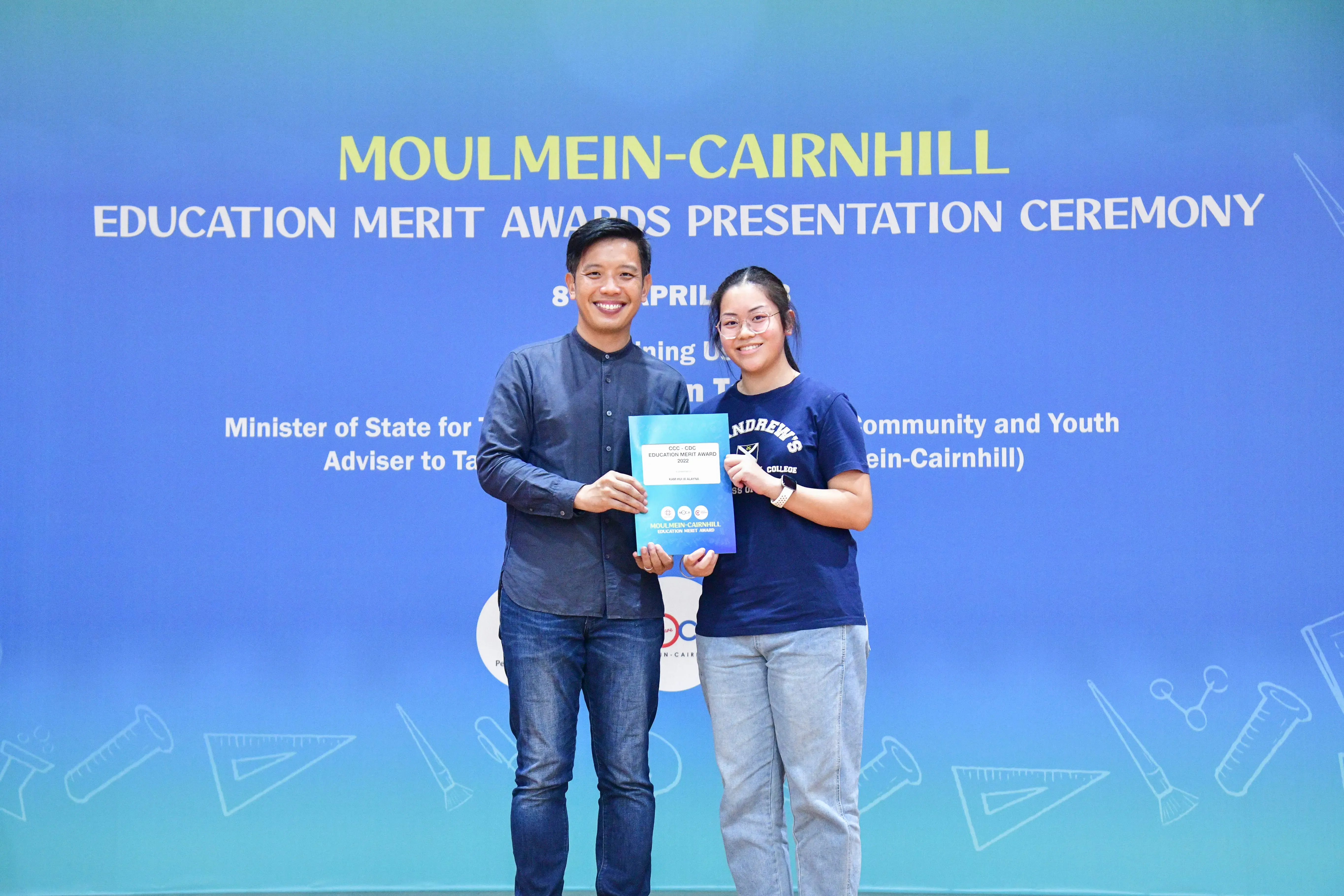 Two smiling people hold an award booklet in front of a Moulmein-Cairnhill Education Merit Awards banner.