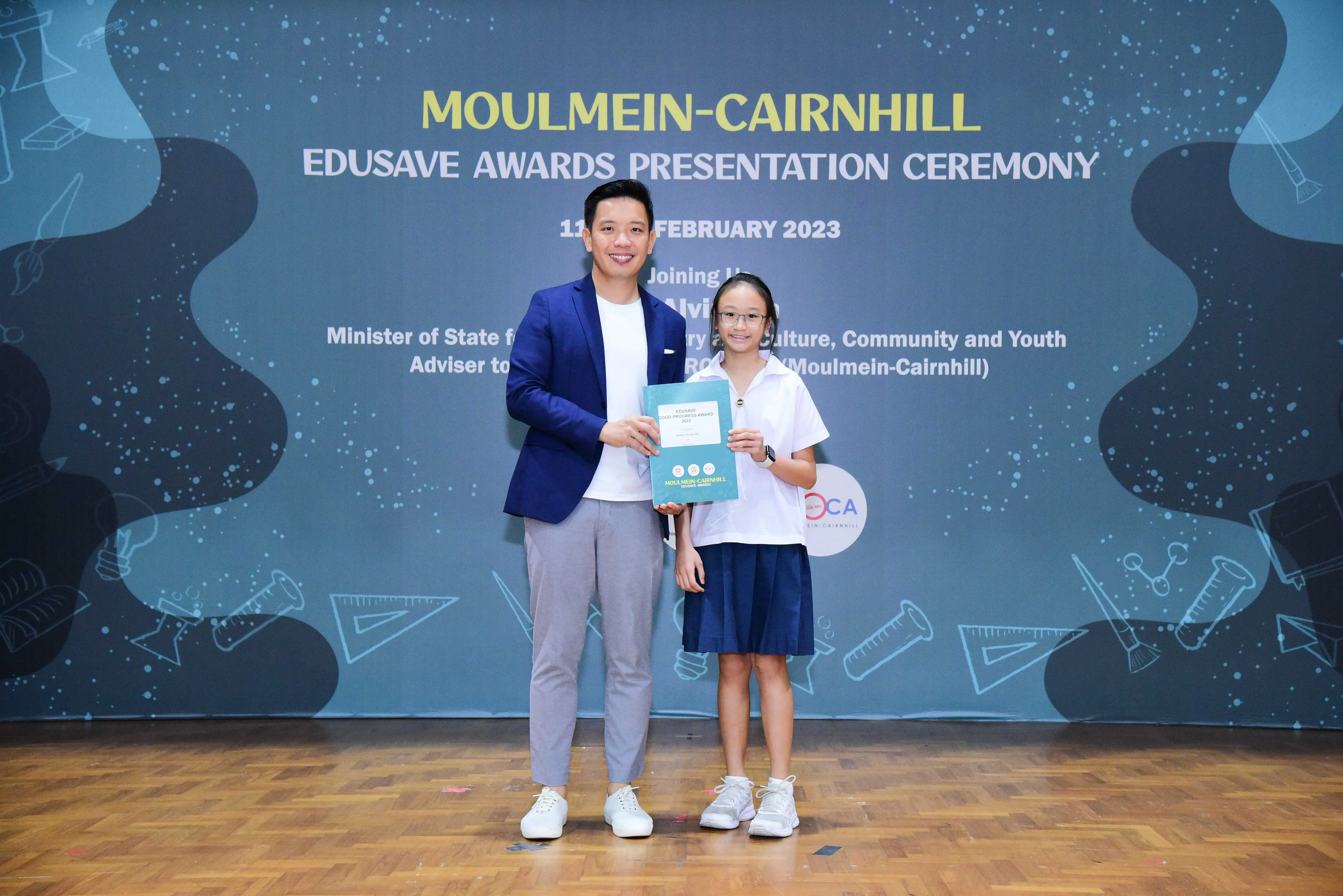 A man in a blue blazer and a young girl in school uniform hold an award at the Moulmein-Cairnhill Edusave Awards Ceremony 2023.