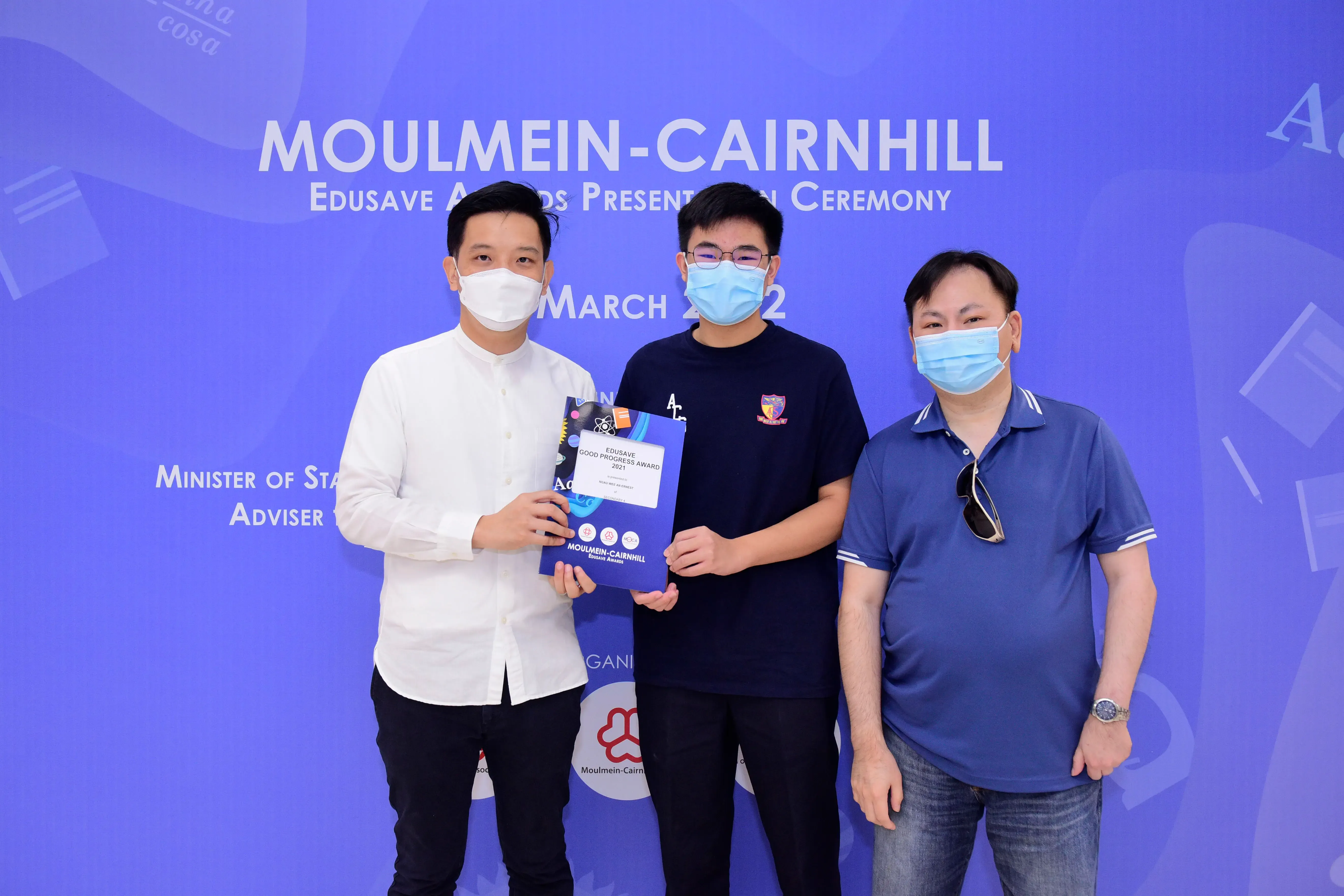 Three masked people pose at the Moulmein-Cairnhill Edusave Awards Presentation Ceremony, with a student holding an award.
