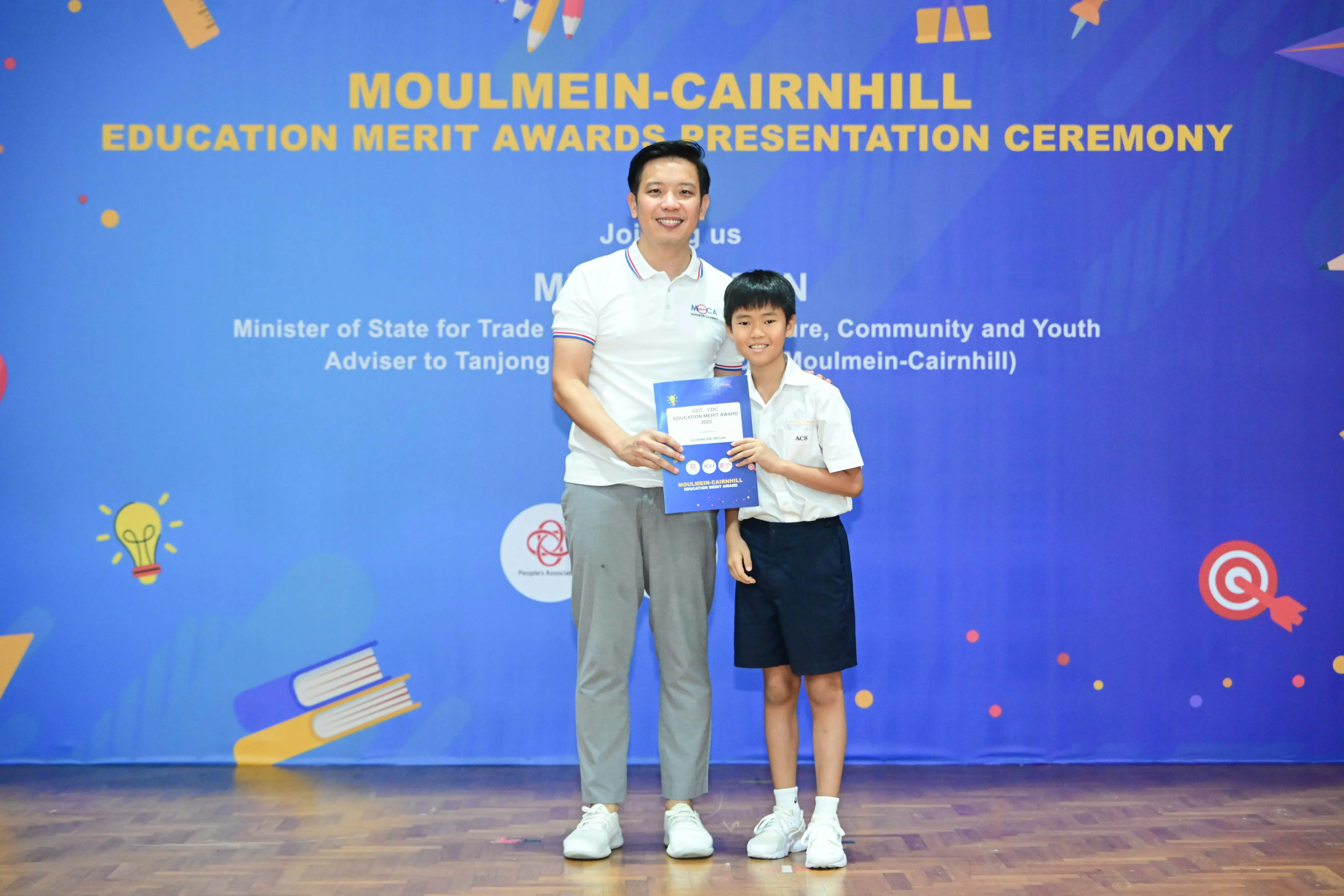 An adult and a young boy hold a certificate together in front of a Moulmein-Cairnhill Education Merit Awards banner.