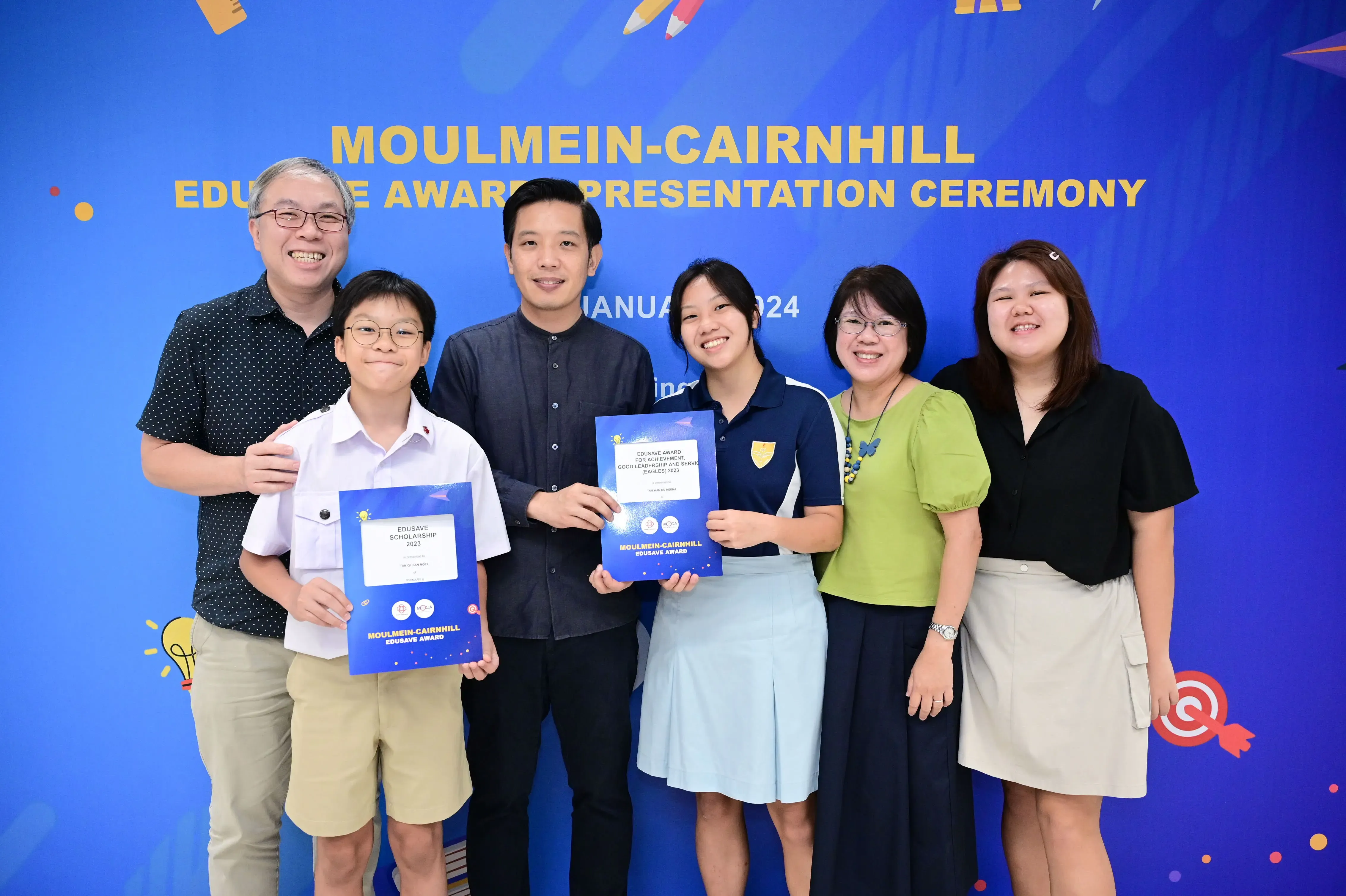 Six people pose smiling at the Moulmein-Cairnhill Edusave Award Presentation Ceremony, two students holding blue award certificates.