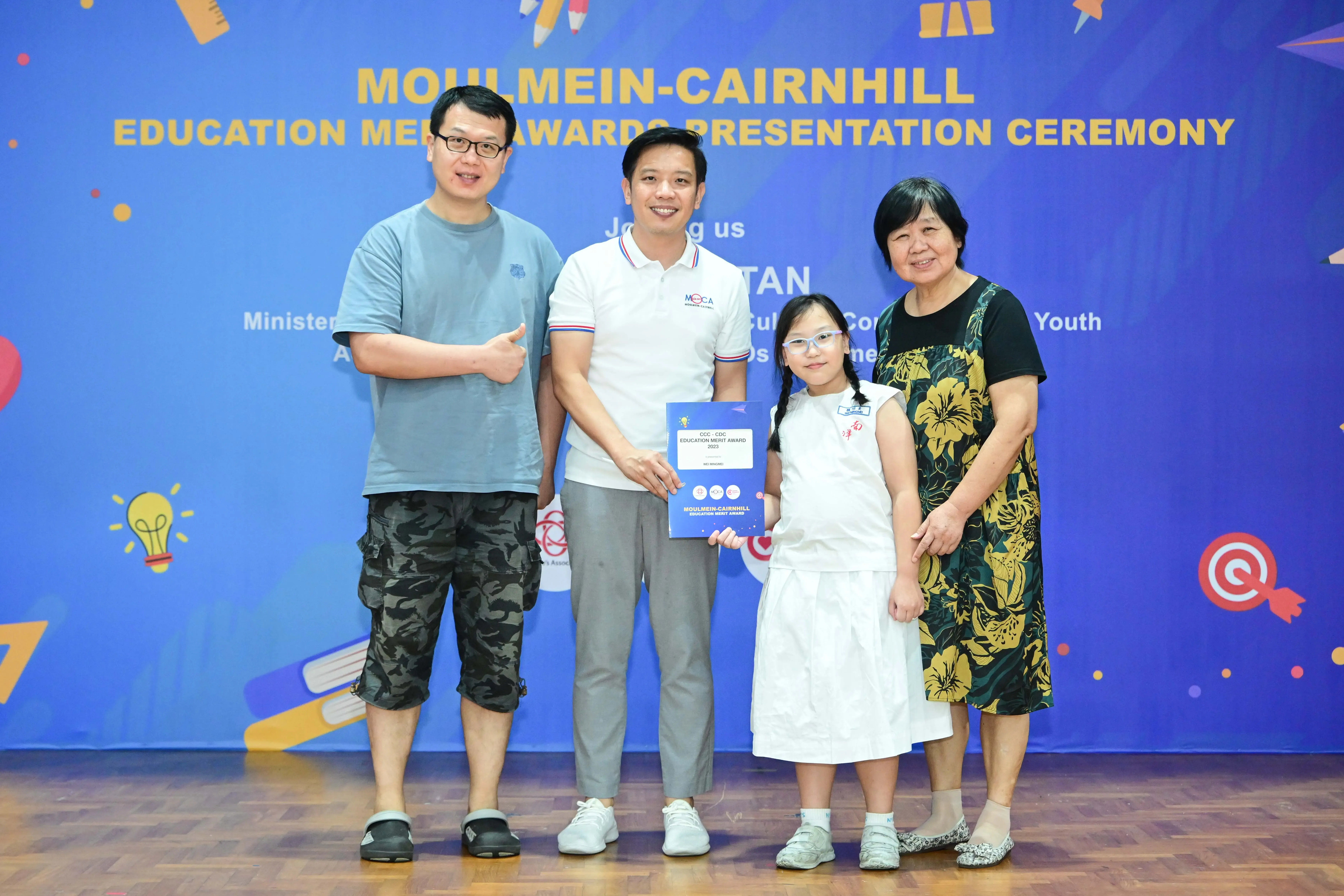 Four people pose at the Moulmein-Cairnhill Education Merit Awards ceremony, including a young girl holding a blue award certificate.