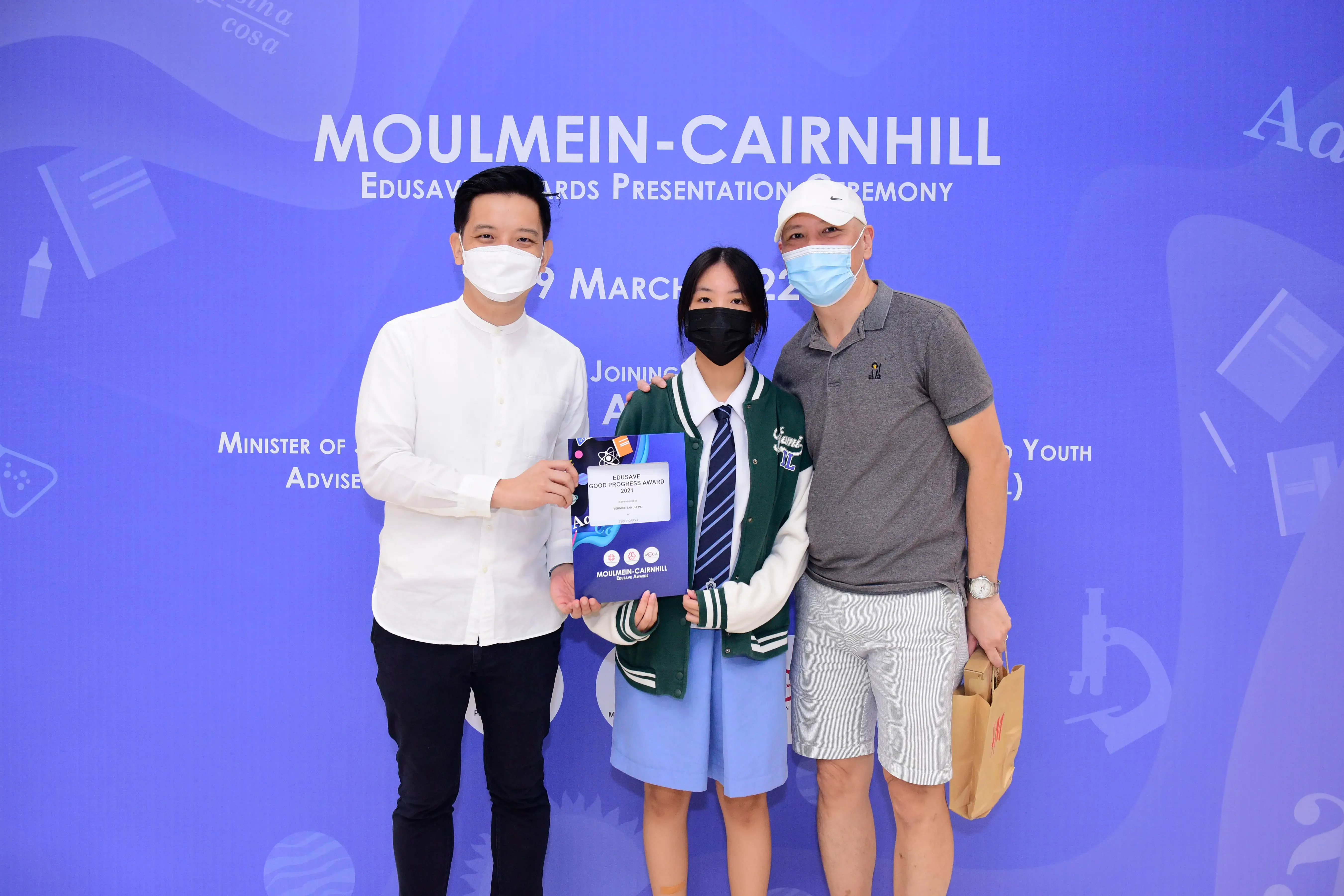 Three masked people pose at the Moulmein-Cairnhill Edusave Awards Presentation Ceremony, with a student holding an award certificate.