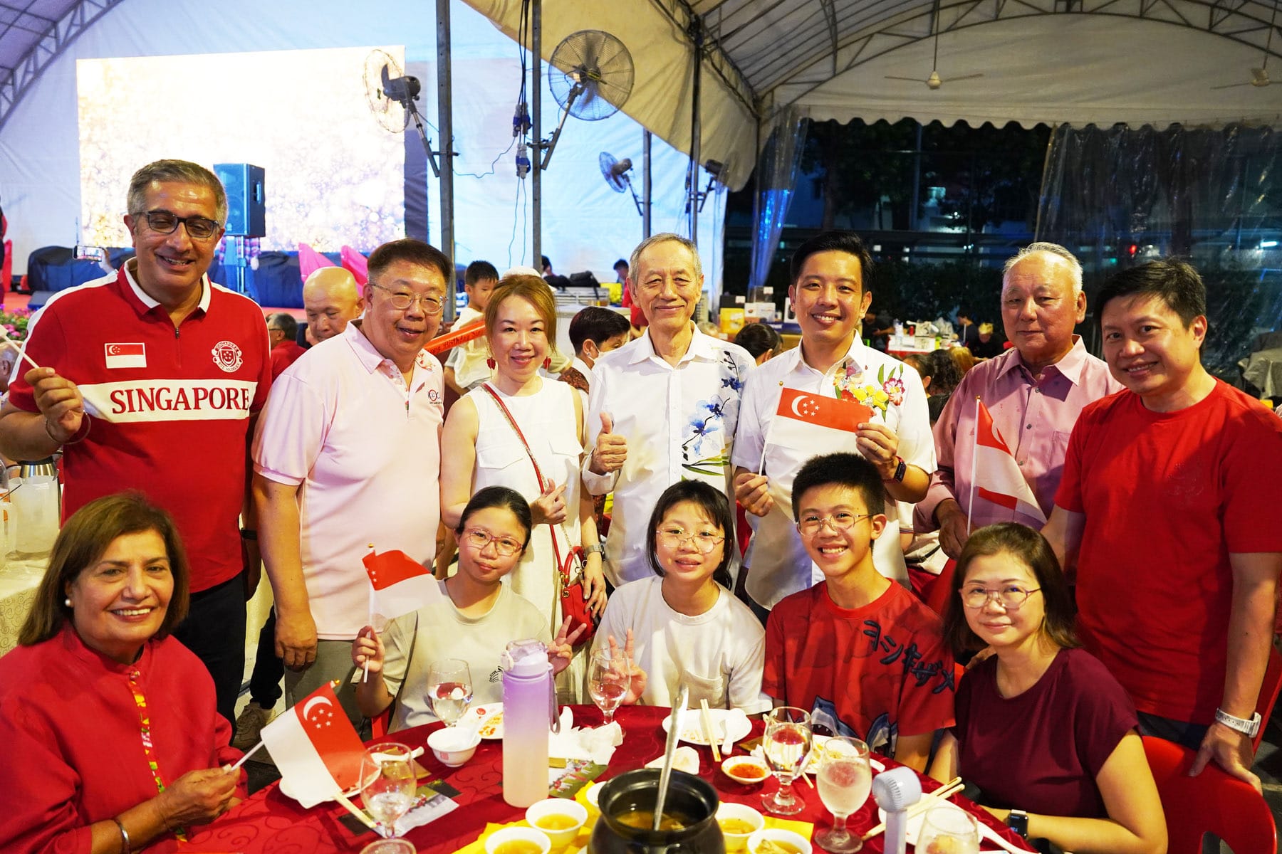 A group of smiling people in red and white clothing hold small Singapore flags at an outdoor dining event under a tent.