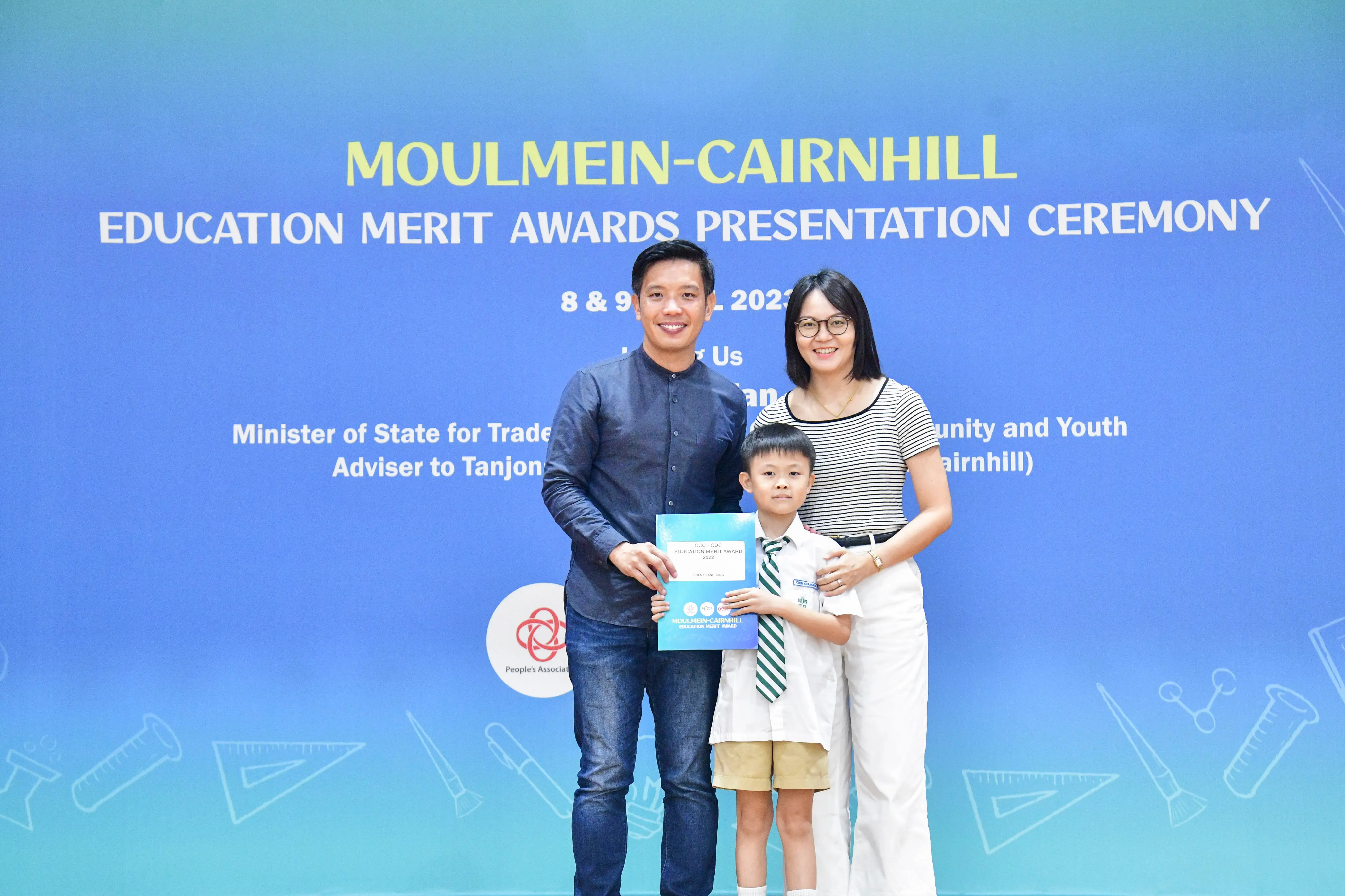 A man, woman, and young boy hold an award certificate in front of a Moulmein-Cairnhill Education Merit Awards banner.