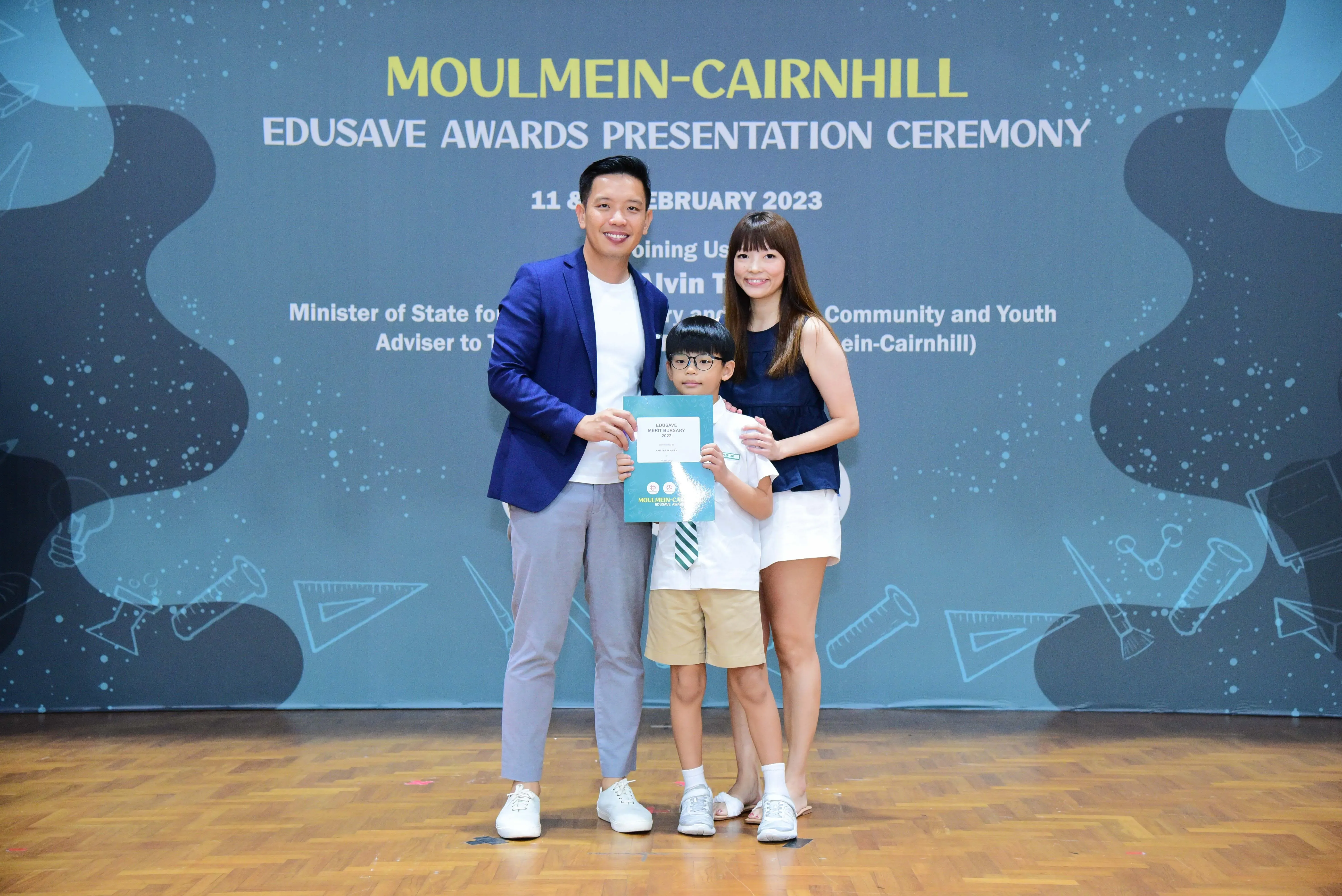 Two adults and a child pose holding a certificate at the Moulmein-Cairnhill Edusave Awards Ceremony, February 2023.