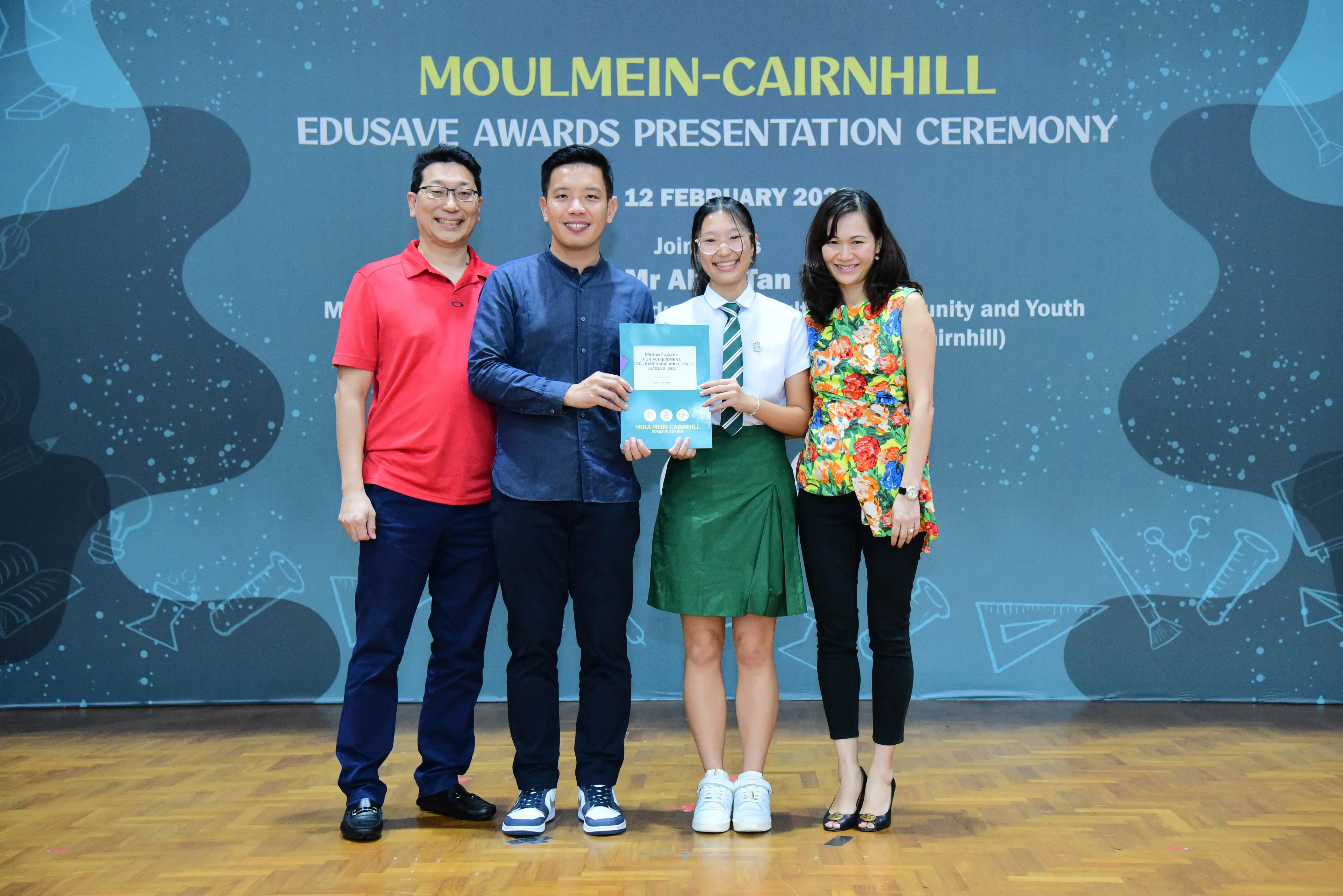 Four people pose holding a certificate at the Moulmein-Cairnhill Edusave Awards Presentation Ceremony backdrop.