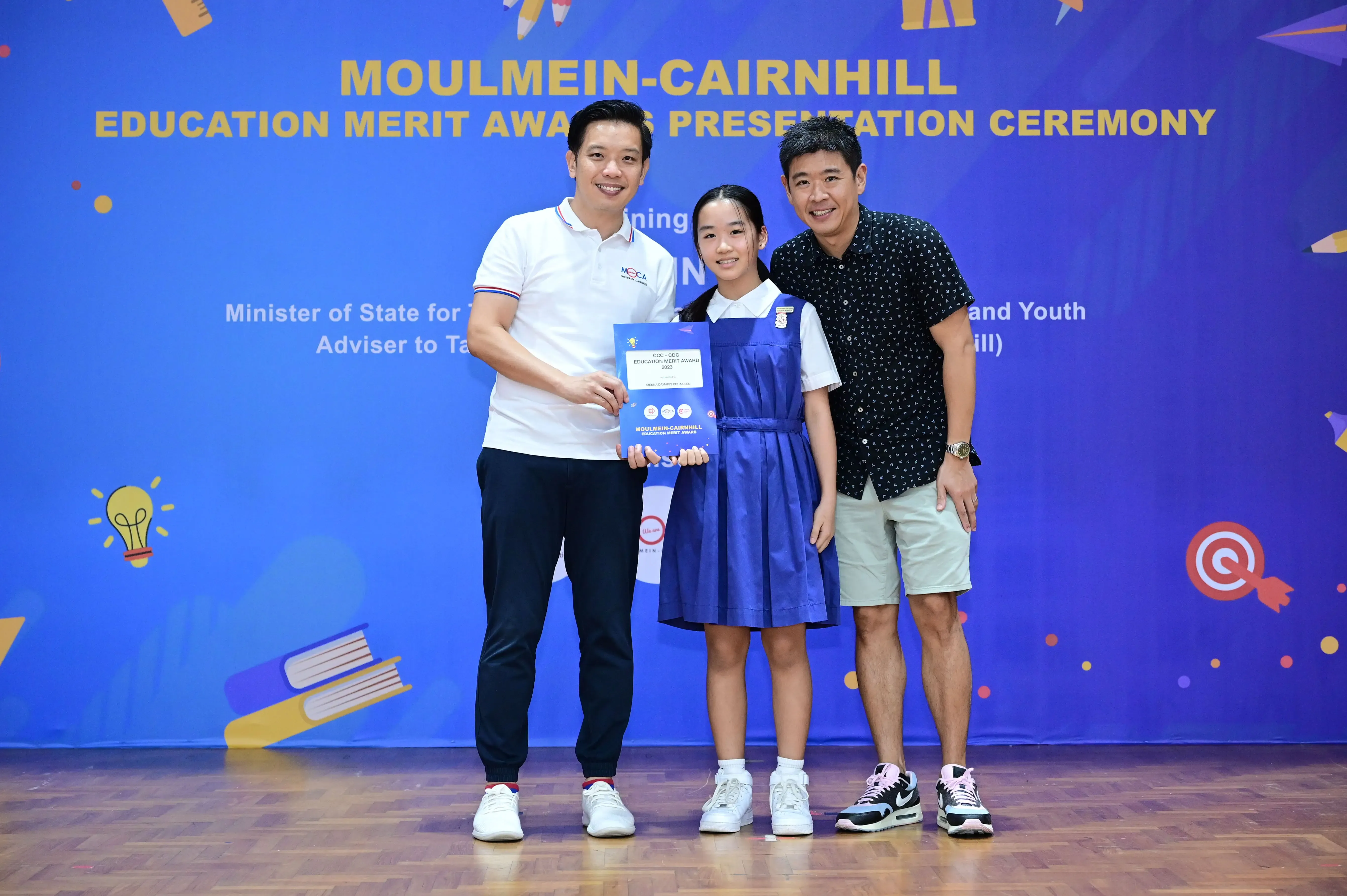 A girl in a blue school uniform holds an award certificate flanked by two men at the Moulmein-Cairnhill Education Merit Awards ceremony.