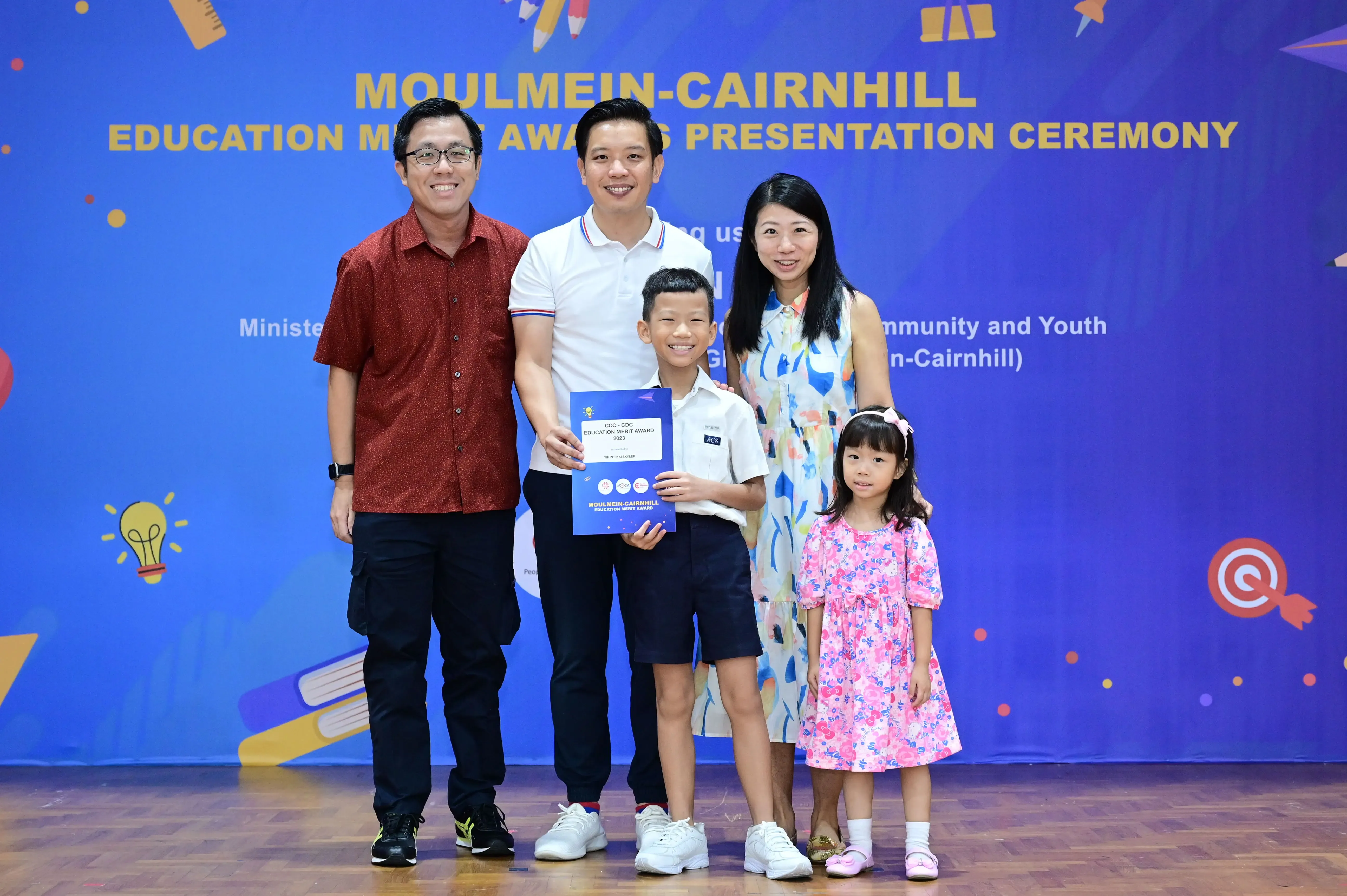 Five people pose at the Moulmein-Cairnhill Education Merit Awards ceremony, with a boy holding a blue certificate.