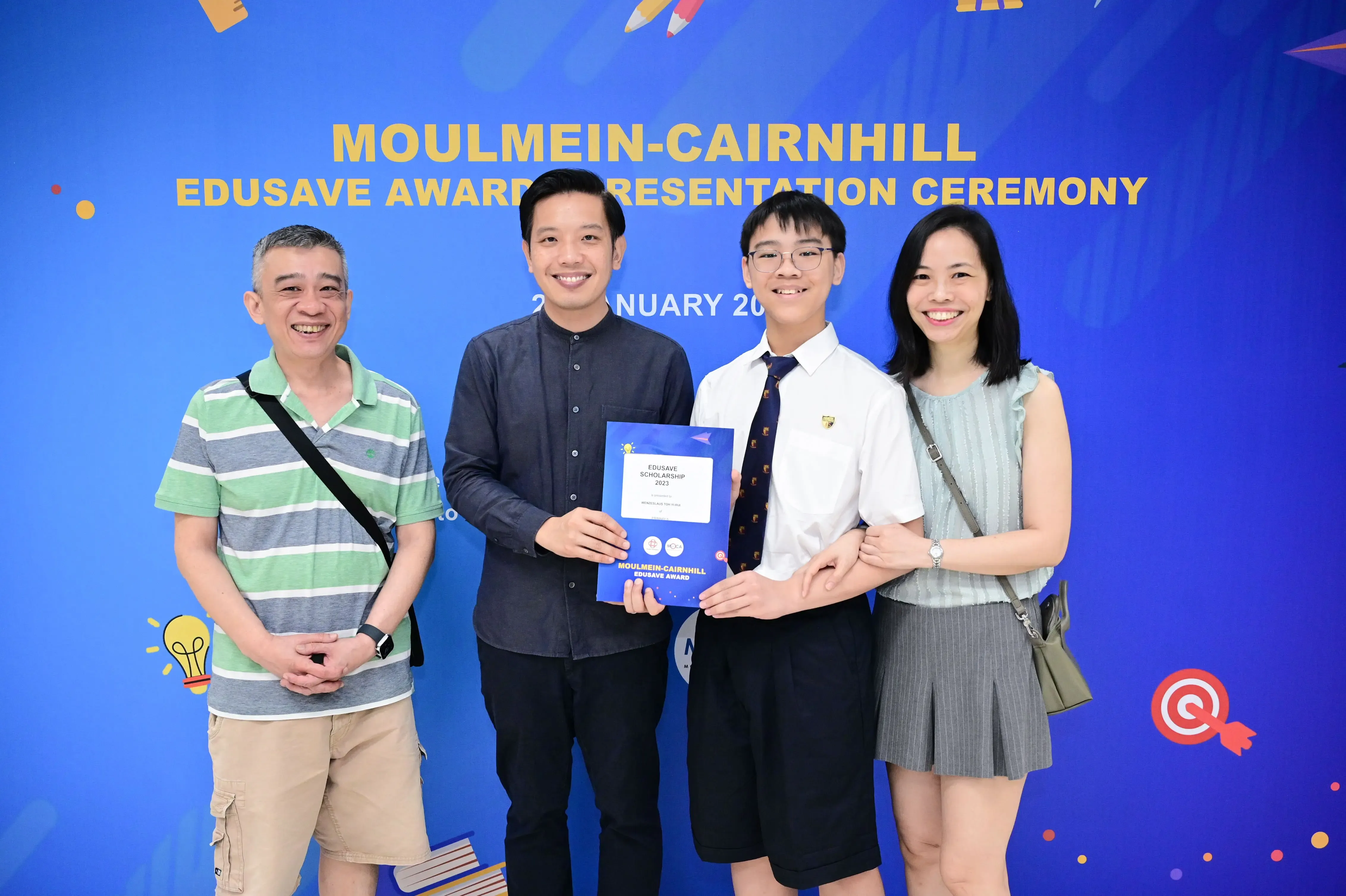 Four people smile in front of a blue Moulmein-Cairnhill Edusave Award Presentation Ceremony banner, holding a certificate.