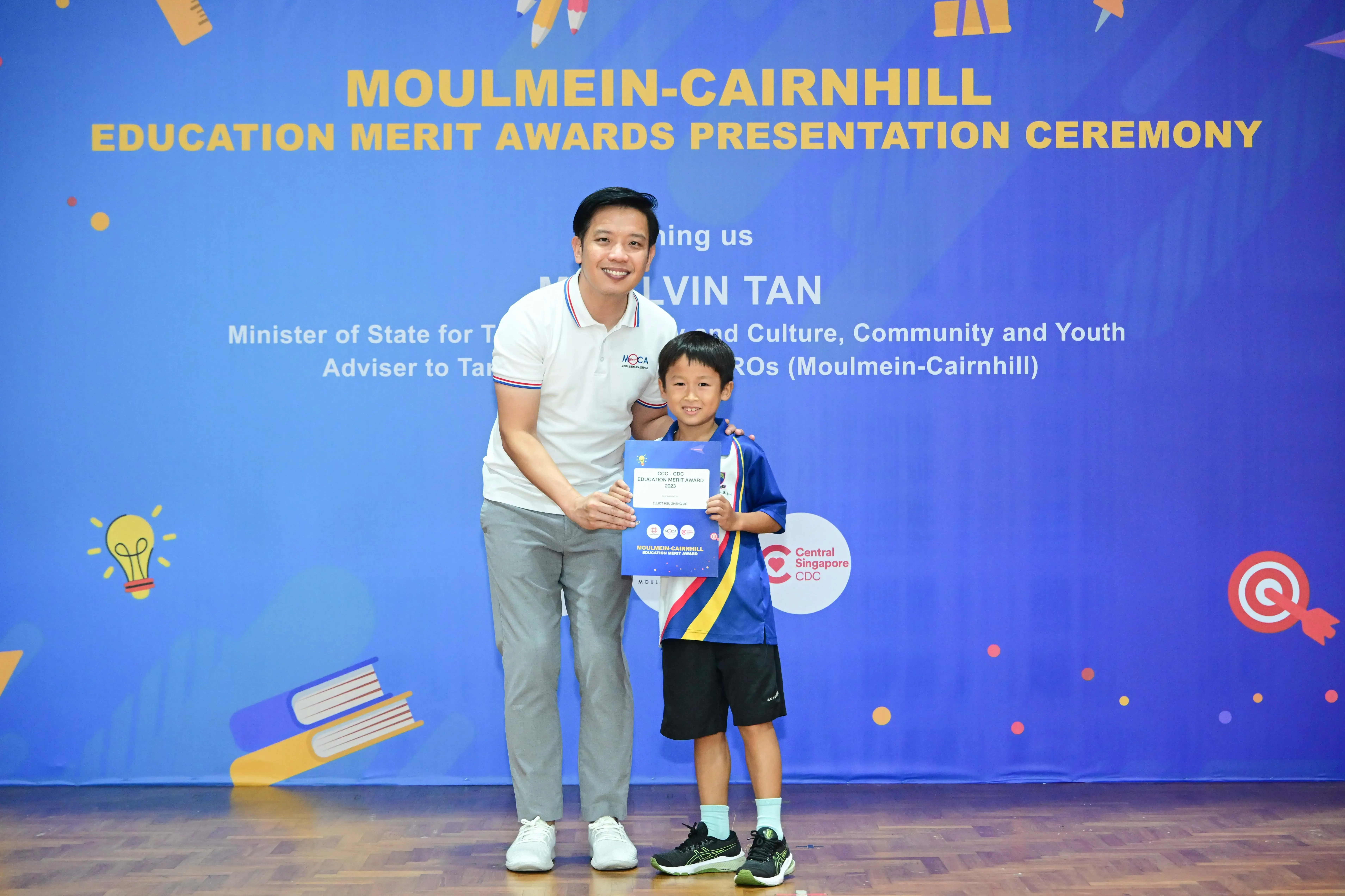 A man and a young boy hold a certificate in front of a Moulmein-Cairnhill Education Merit Awards banner.