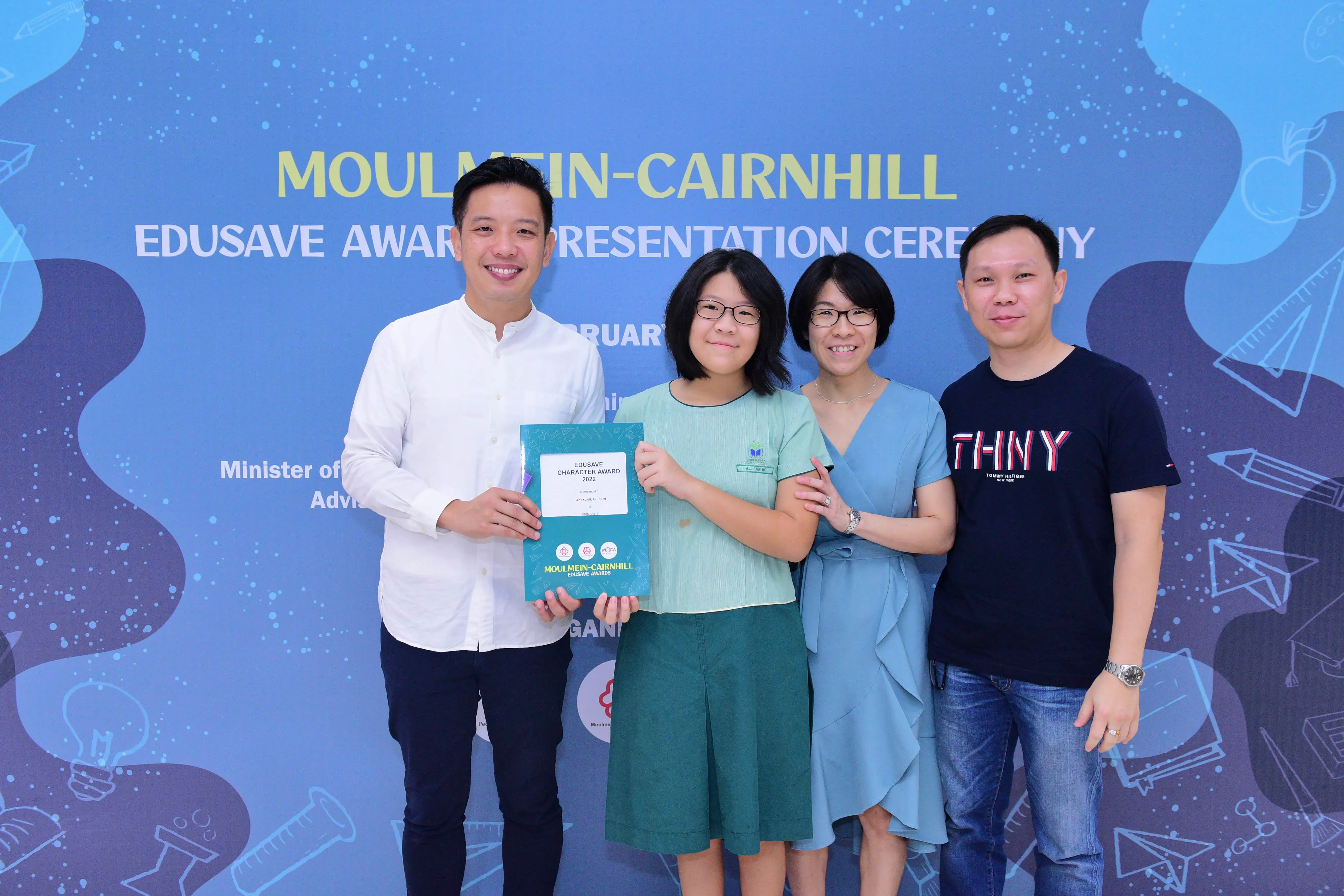 Four people pose smiling at the Moulmein-Cairnhill Edusave Award Ceremony, with a girl holding a certificate.