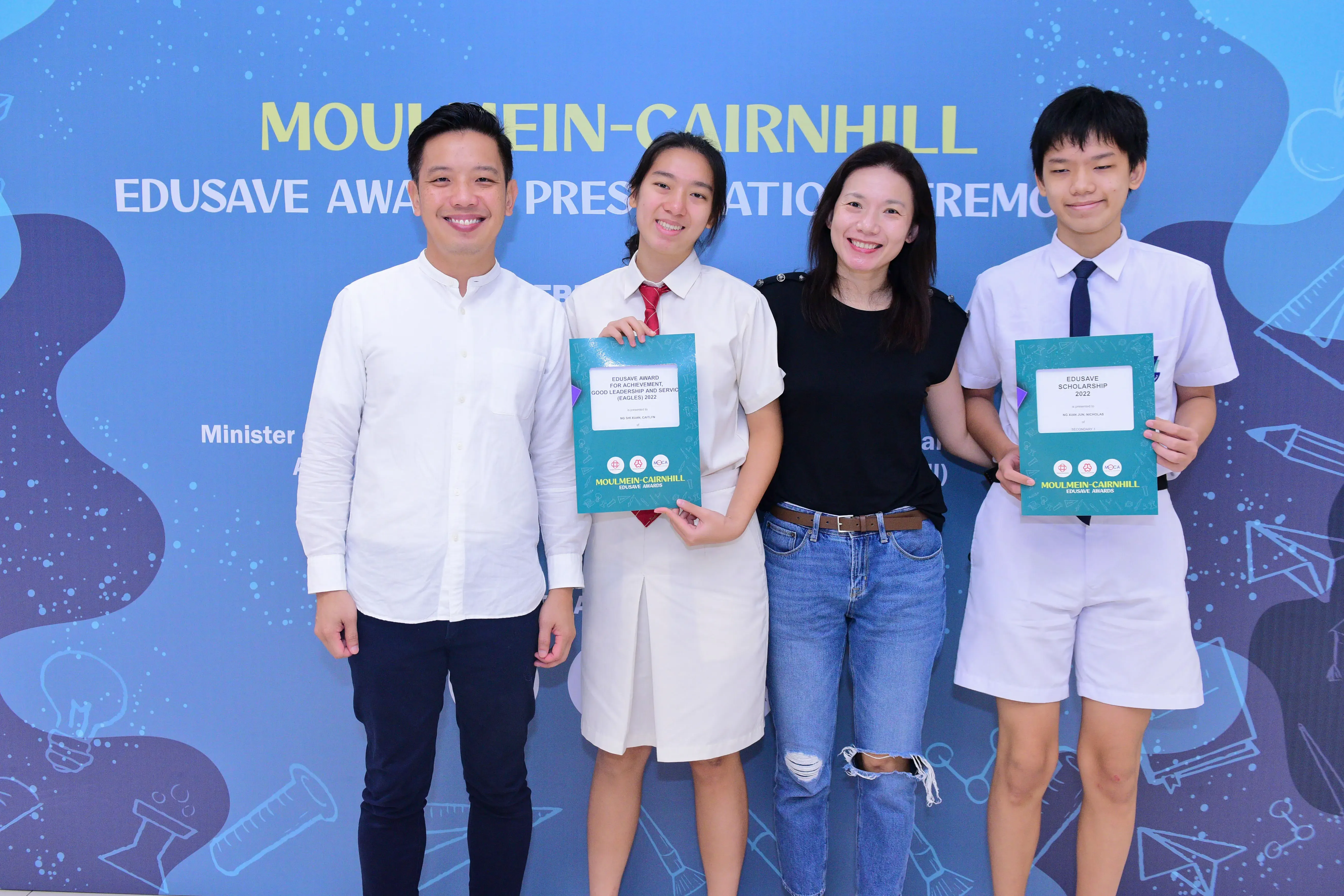 Four smiling people pose before a Moulmein-Cairnhill Edusave Award Presentation Ceremony banner, two holding award certificates.