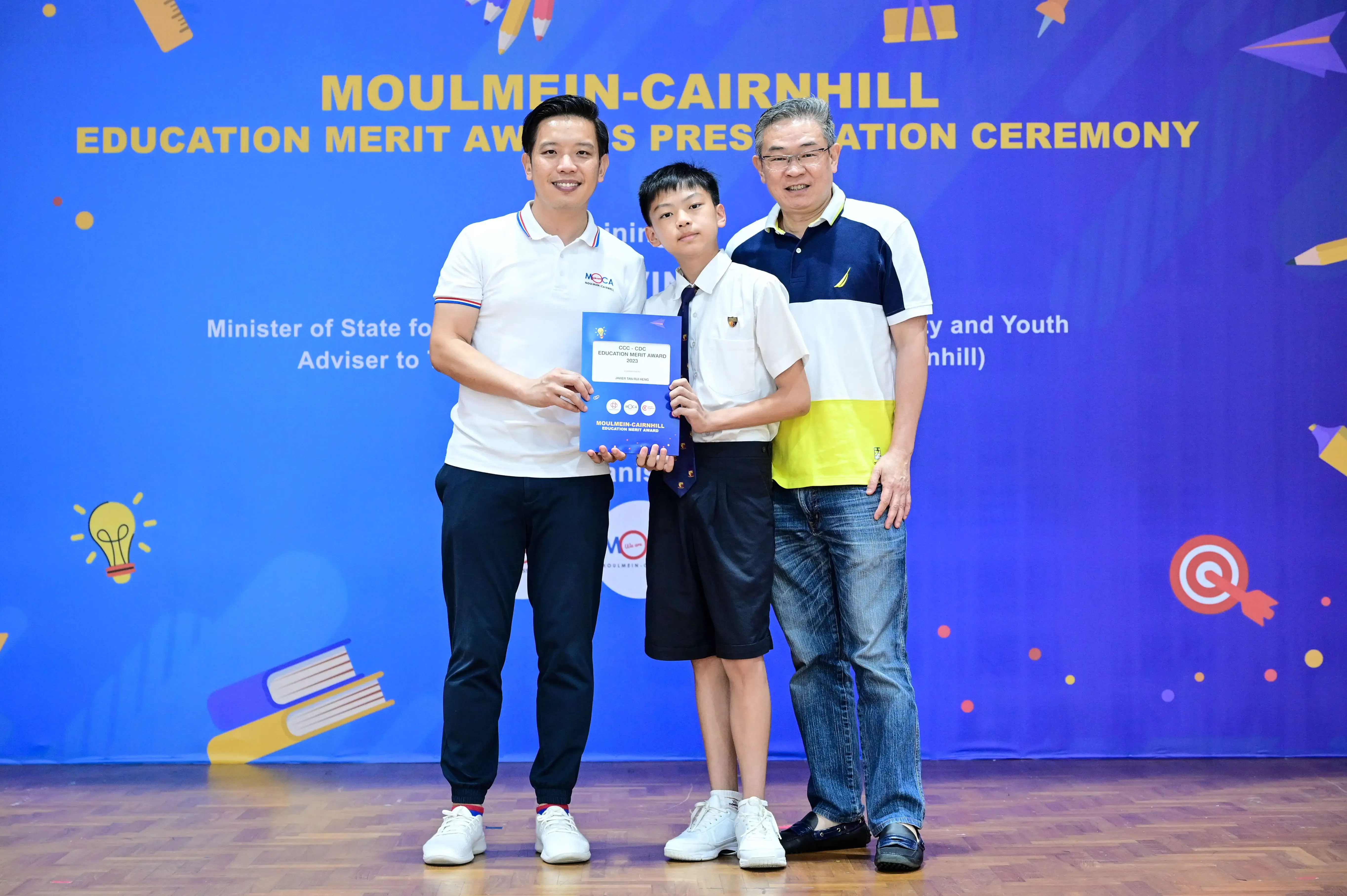 Two men and a boy in school uniform hold a certificate at the Moulmein-Cairnhill Education Merit Awards ceremony.