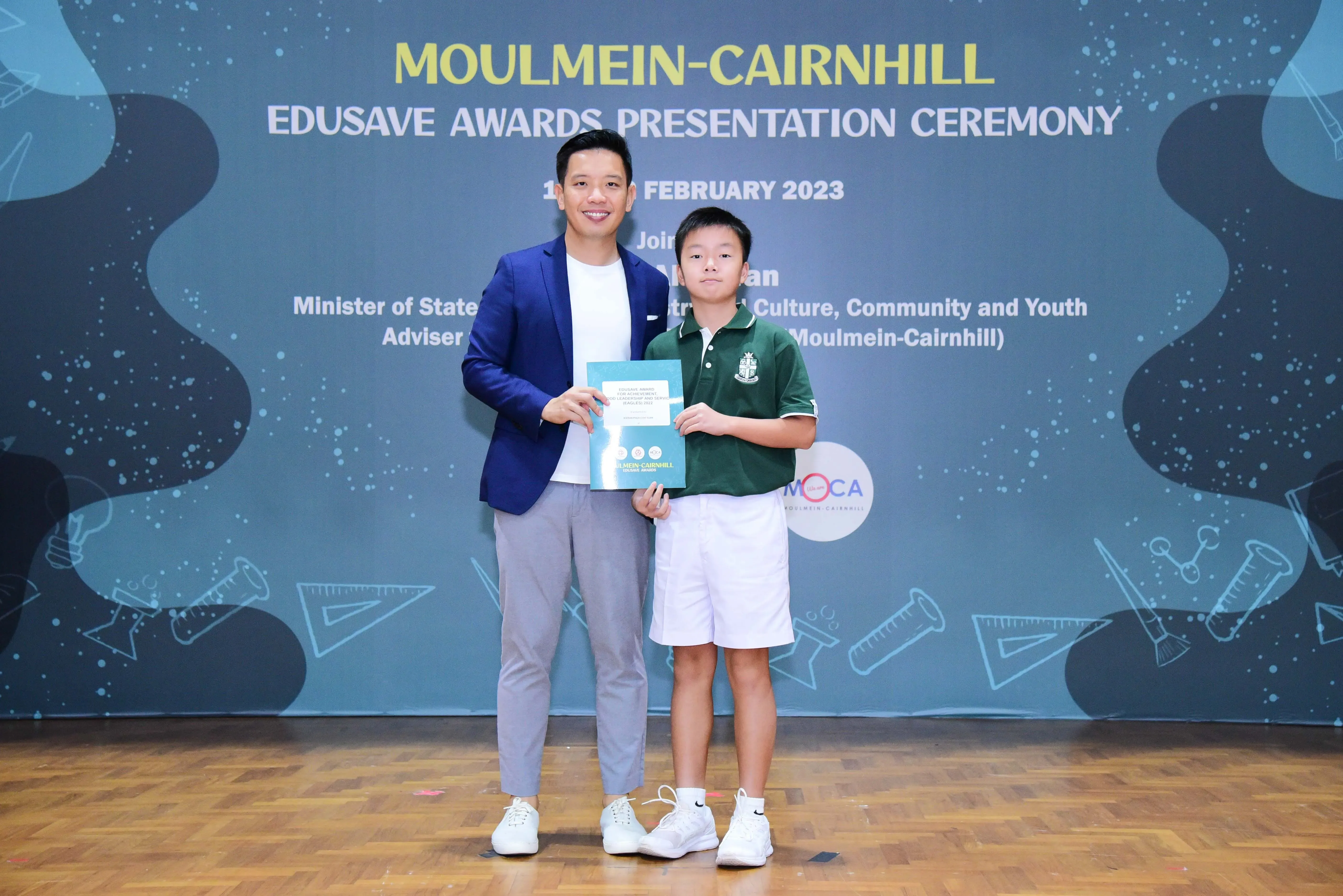 A man in a blue blazer and a young boy in school uniform pose holding a certificate at the Moulmein-Cairnhill Edusave Awards 2023.