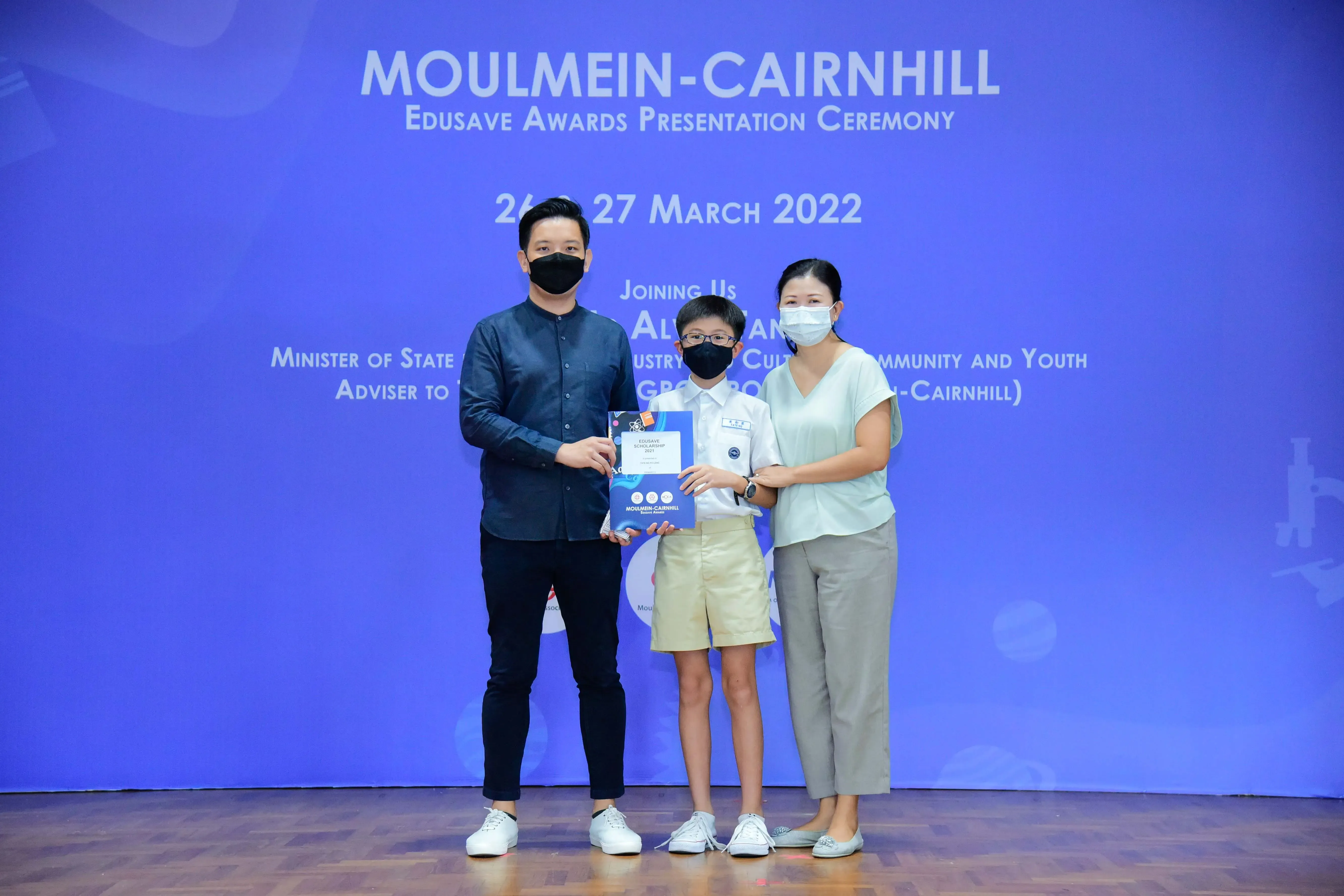 Two masked adults and a masked boy hold an award at the Moulmein-Cairnhill Edusave Awards Ceremony, March 2022.