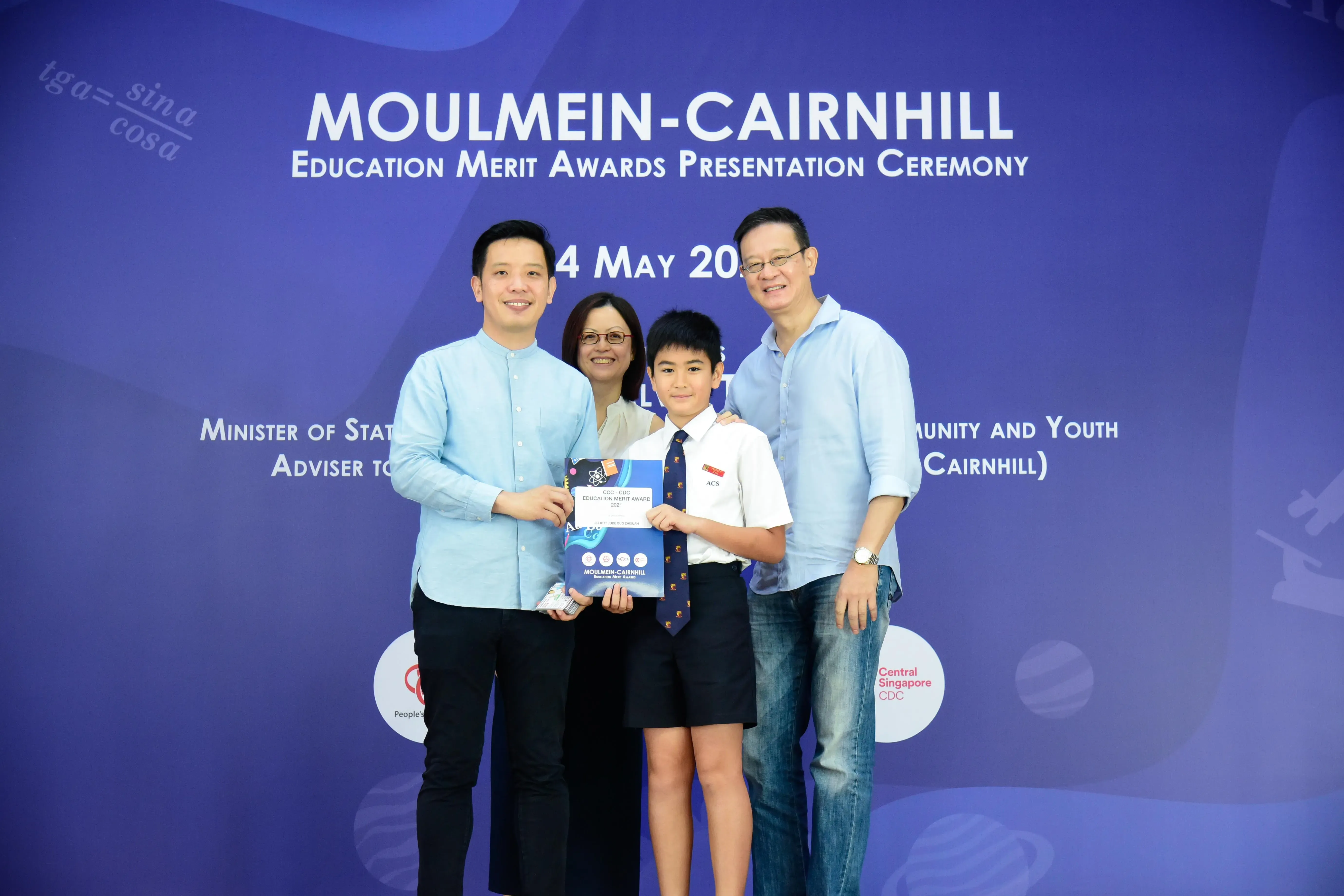 Four people pose with an award at the Moulmein-Cairnhill Education Merit Awards Presentation Ceremony backdrop.