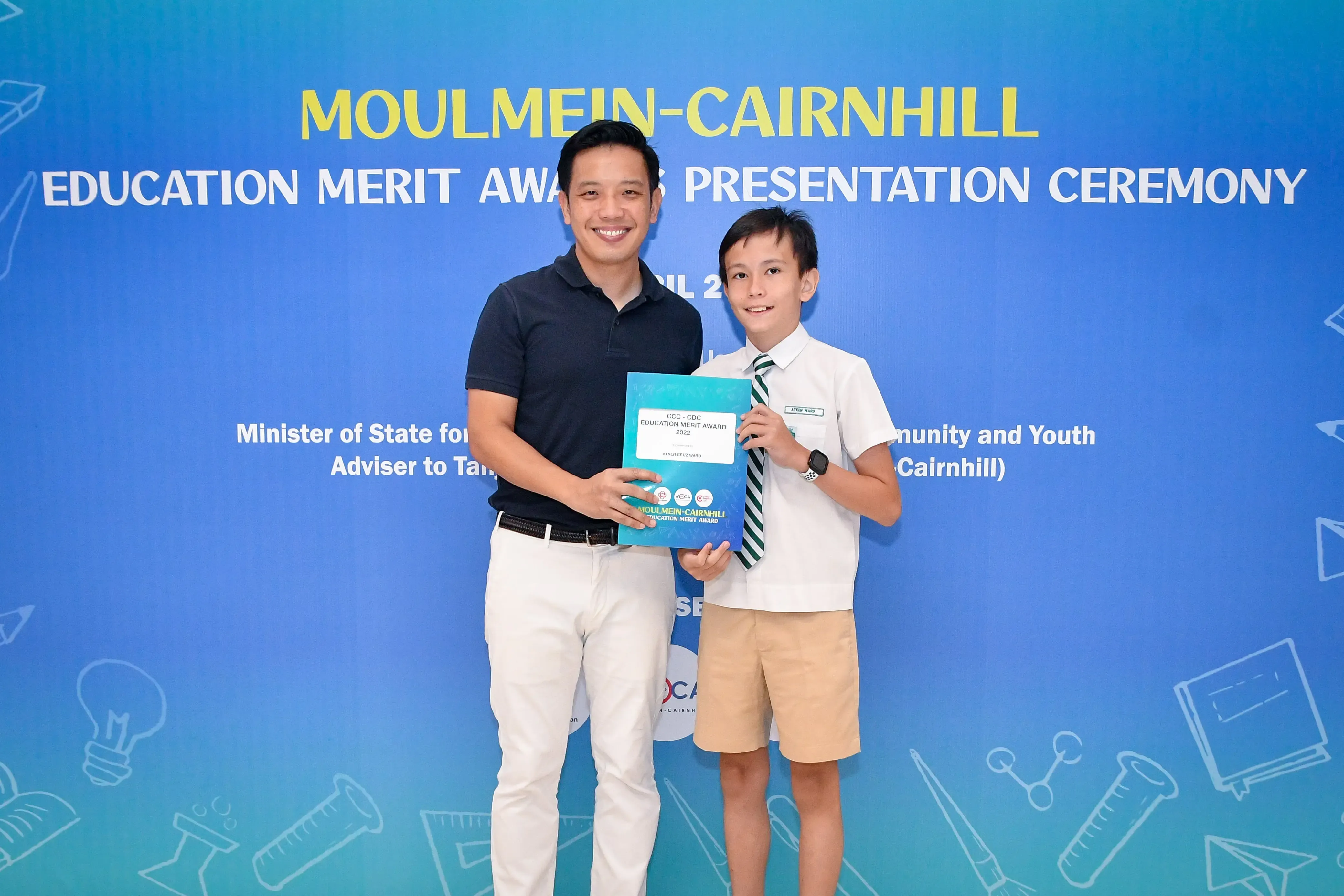 A man and a young boy in school uniform hold an award certificate at the Moulmein-Cairnhill Education Merit Awards ceremony.