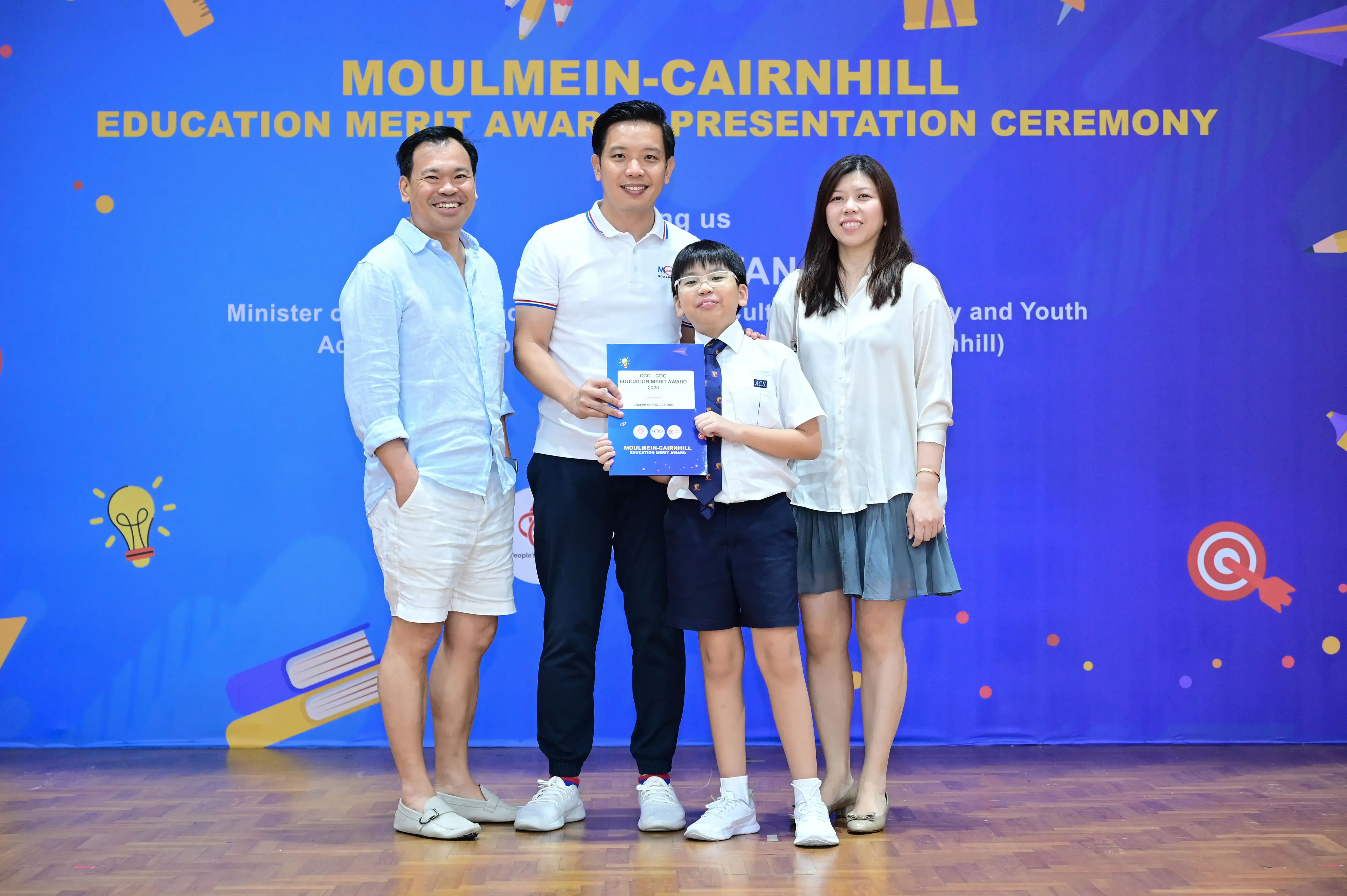 Three adults and a young boy in school uniform hold a certificate at the Moulmein-Cairnhill Education Merit Award ceremony.