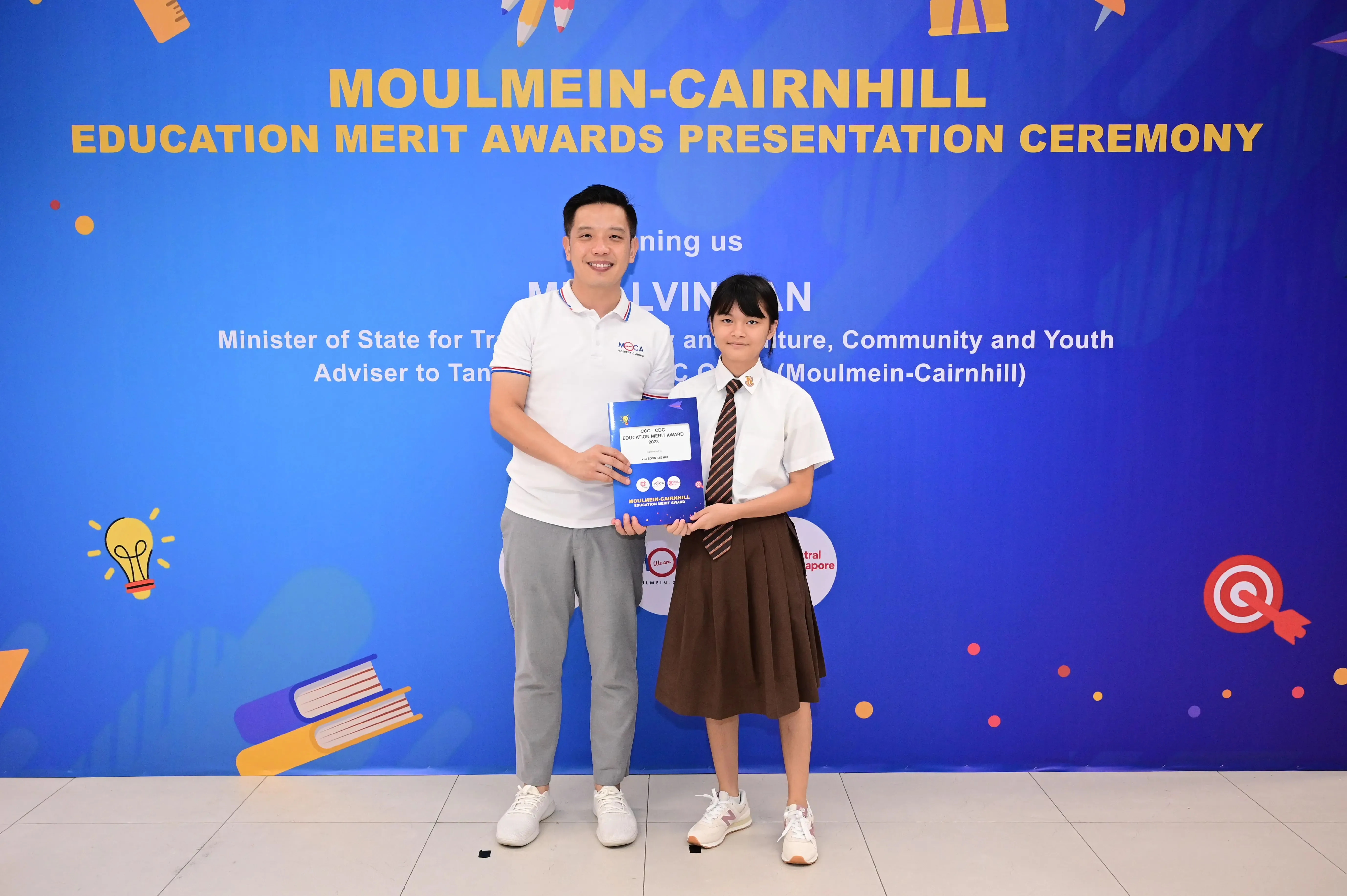 A man and a uniformed schoolgirl hold an award certificate at the Moulmein-Cairnhill Education Merit Awards ceremony.