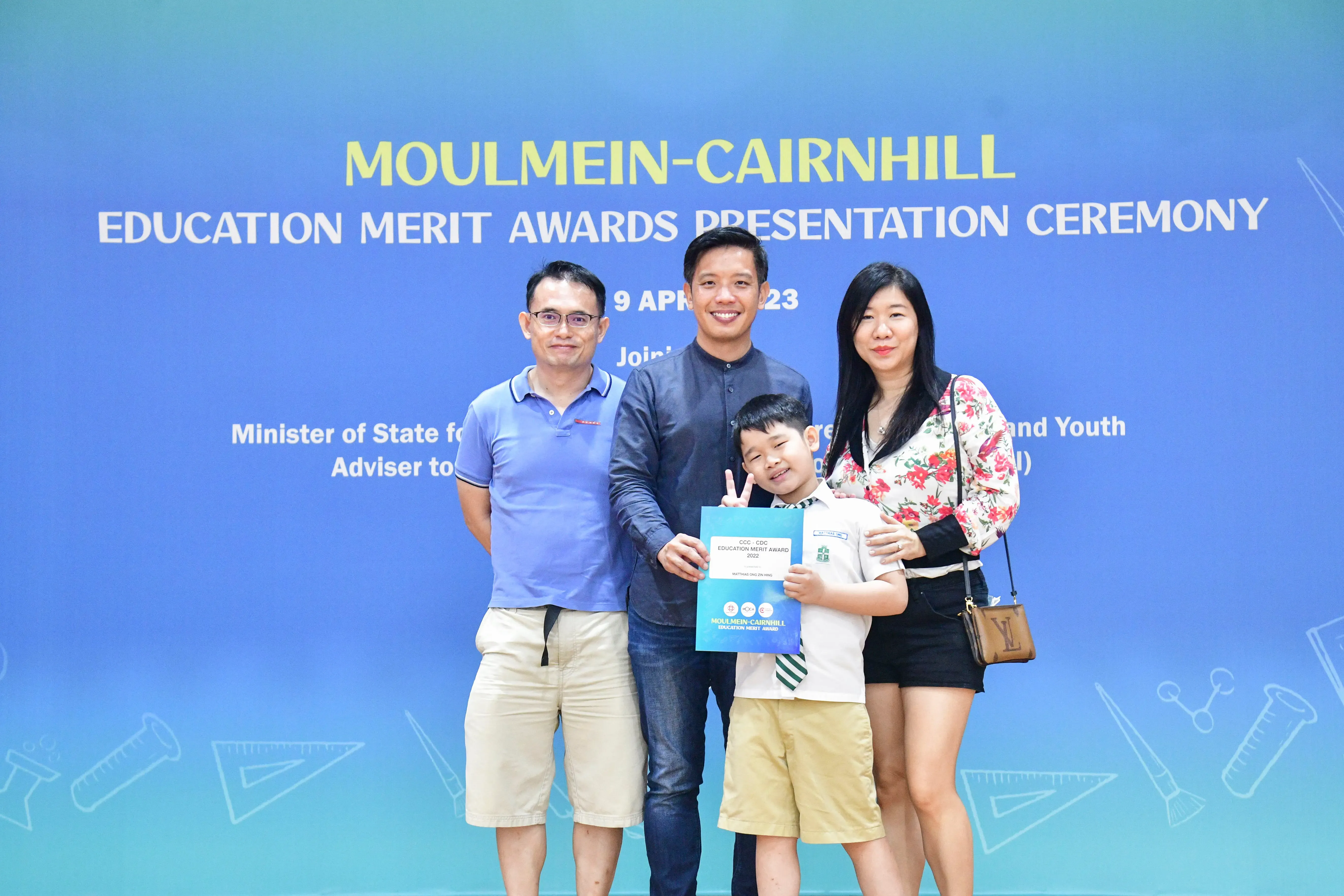 Three adults and a young boy pose at the Moulmein-Cairnhill Education Merit Awards Presentation Ceremony, boy holds a certificate.