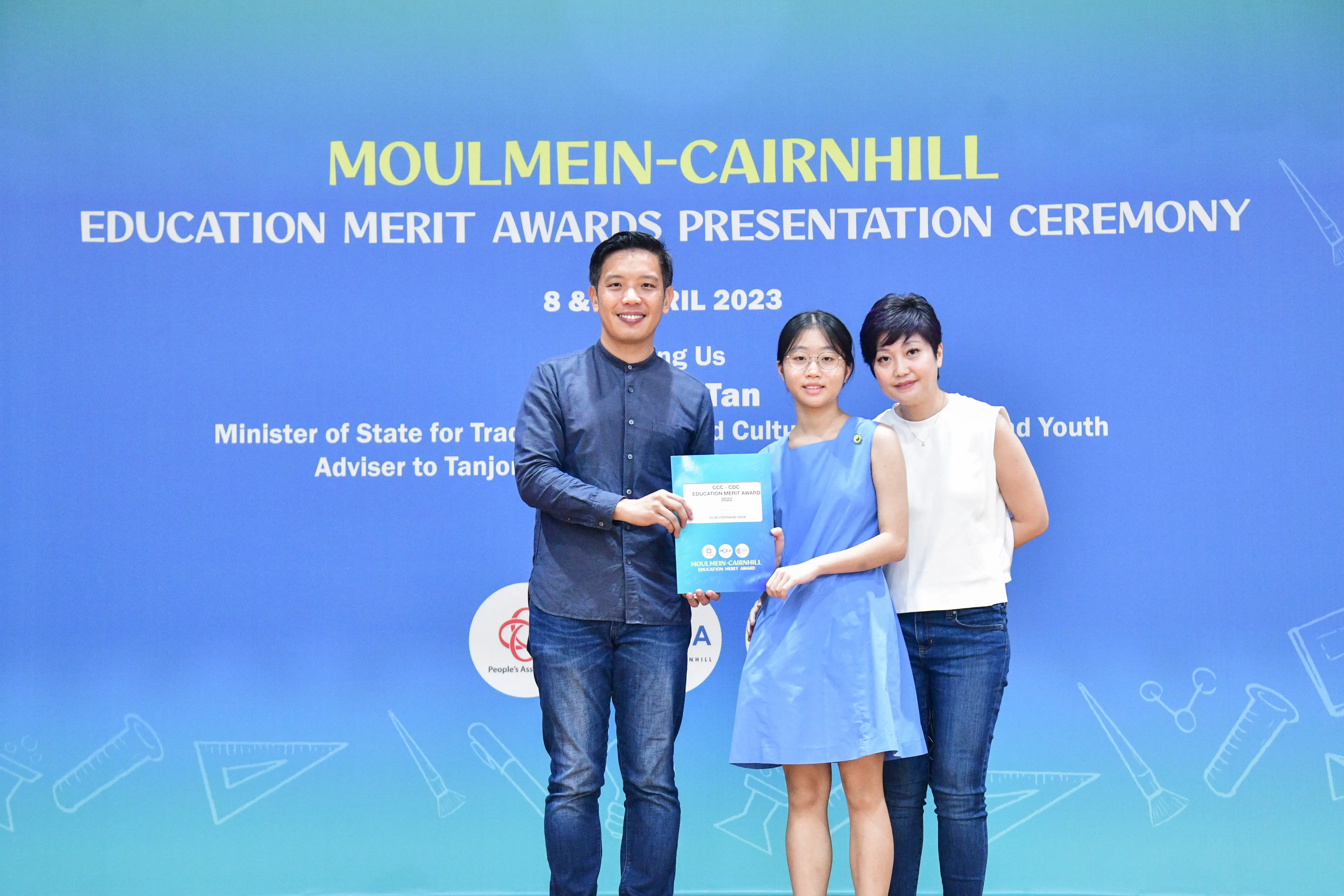 Three people pose holding an award folder at the Moulmein-Cairnhill Education Merit Awards Presentation Ceremony, April 2023.
