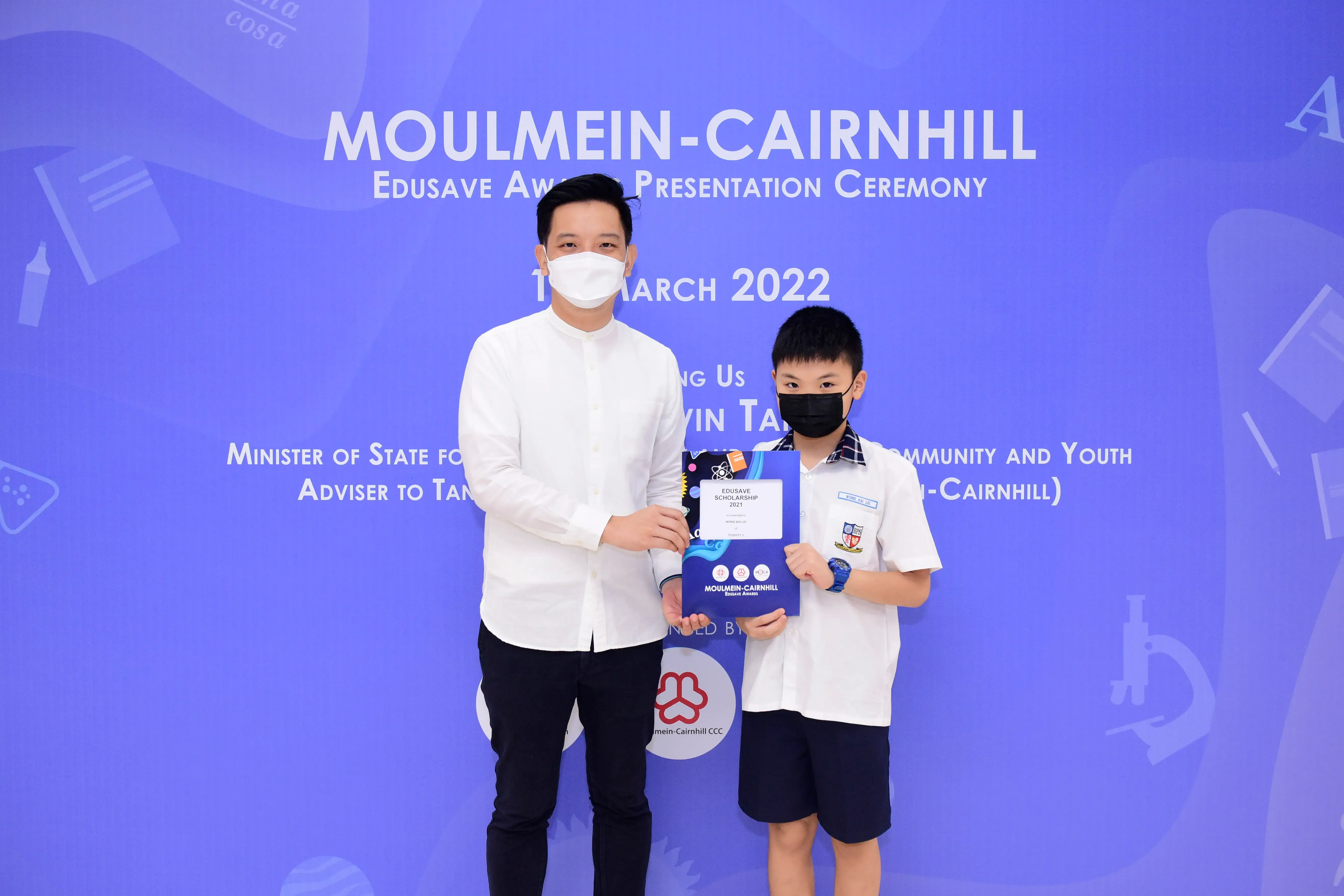 An adult and a young boy hold an Edusave Scholarship certificate at the Moulmein-Cairnhill Award Ceremony, March 2022.