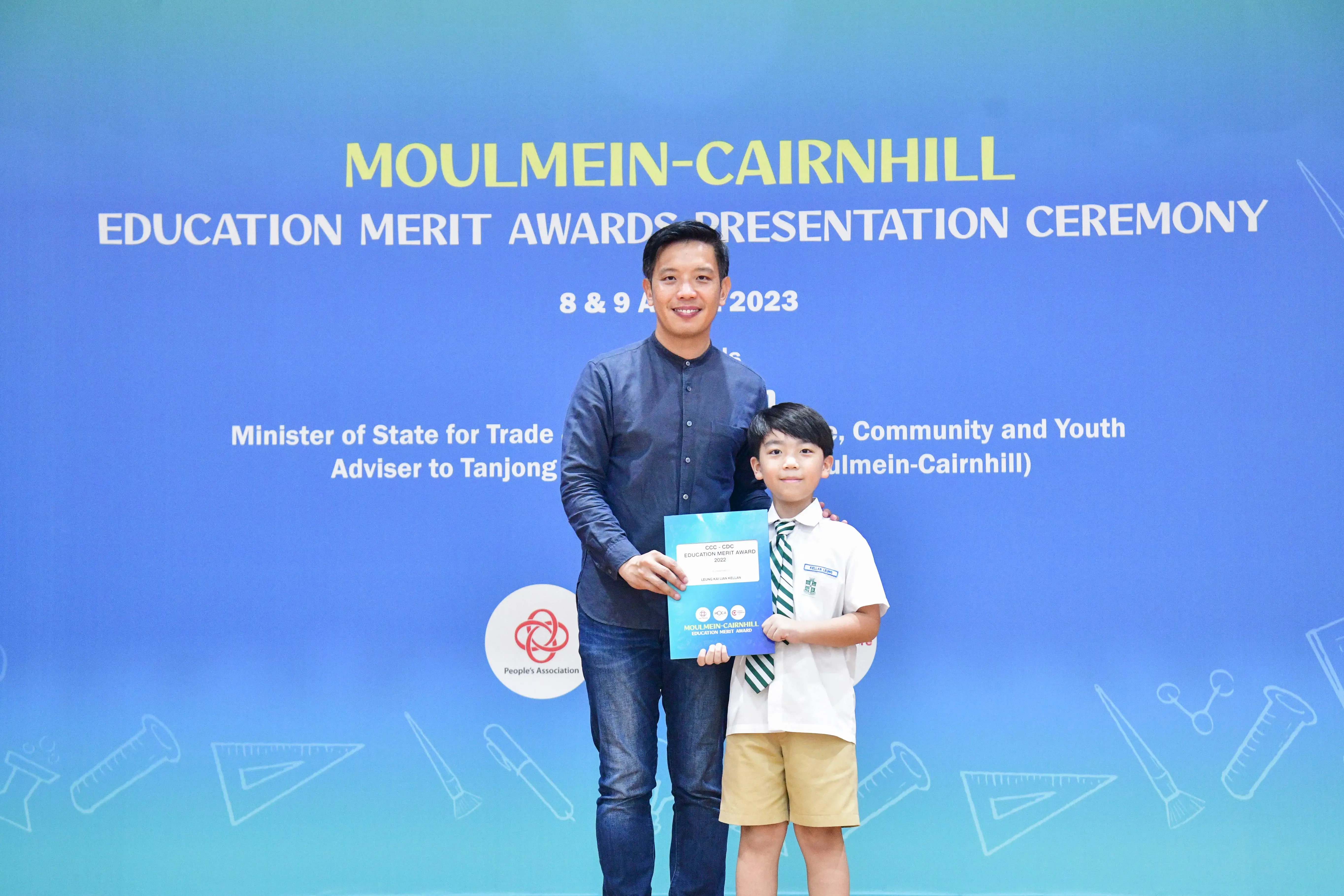 A man and a young boy holding a certificate stand before a Moulmein-Cairnhill Education Merit Awards banner dated 2023.