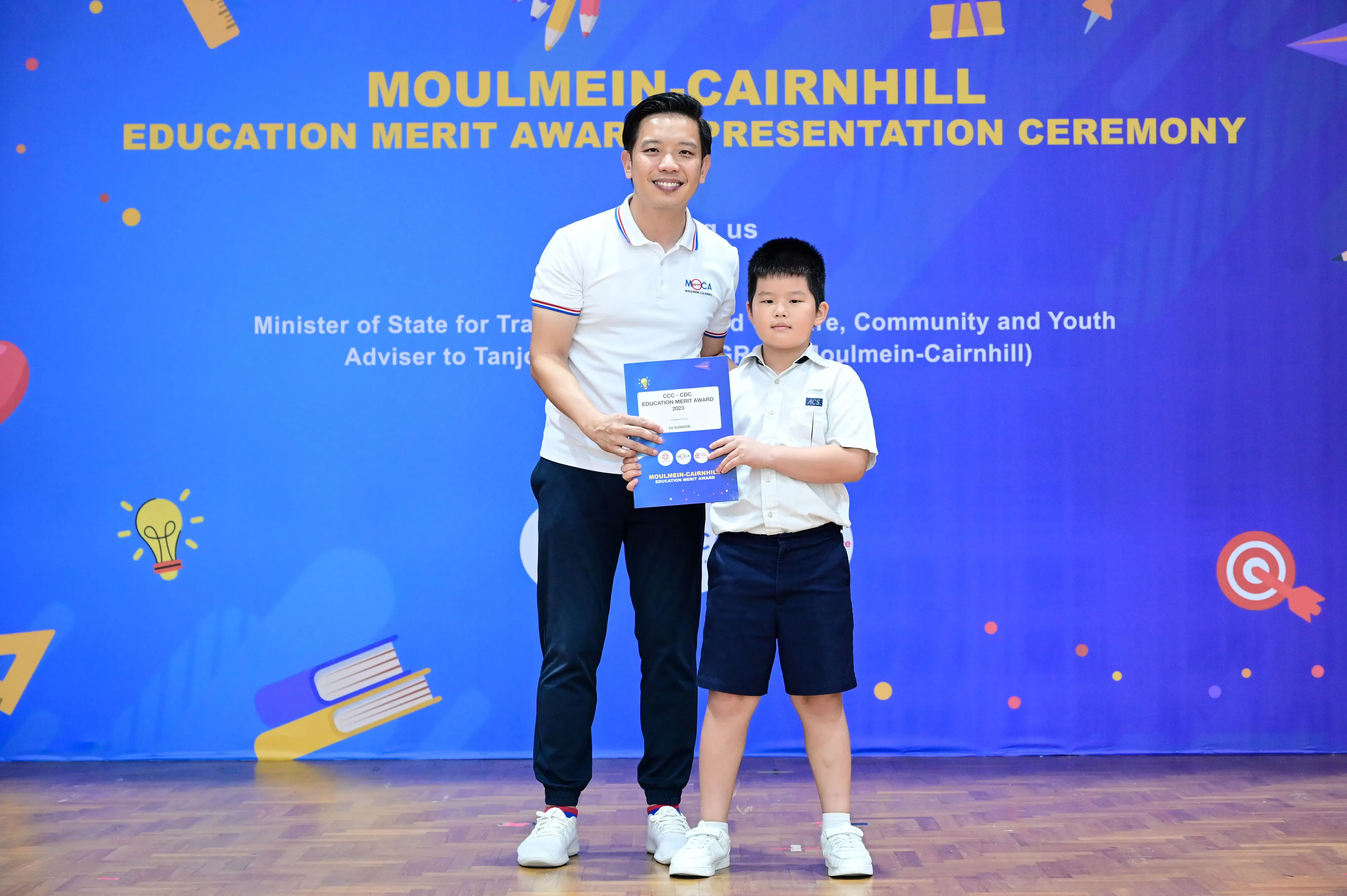An adult and a young boy hold a certificate together at the Moulmein-Cairnhill Education Merit Award Ceremony.