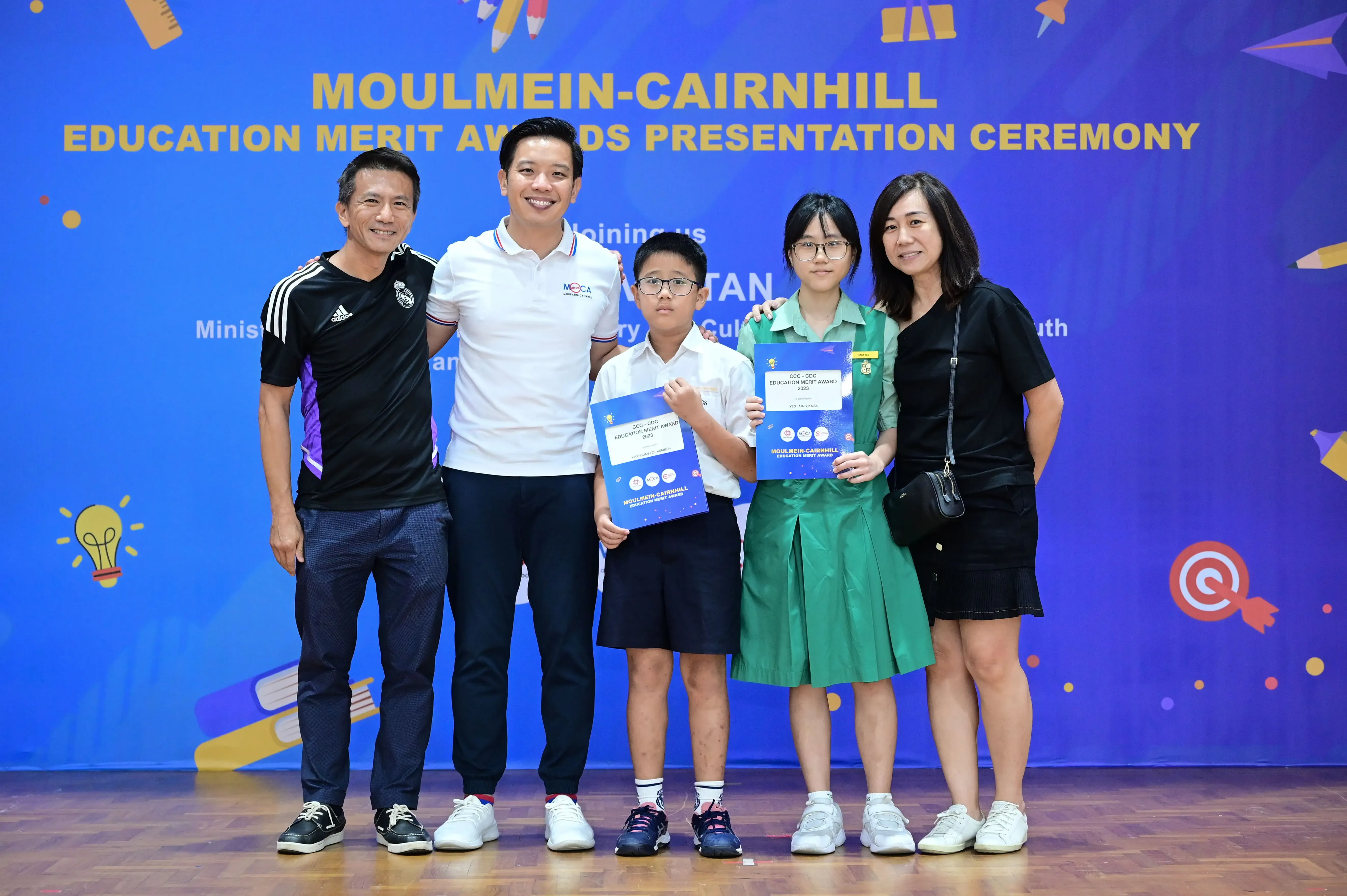 Five people pose in front of a blue Moulmein-Cairnhill Education Merit Awards banner, two children holding award certificates.