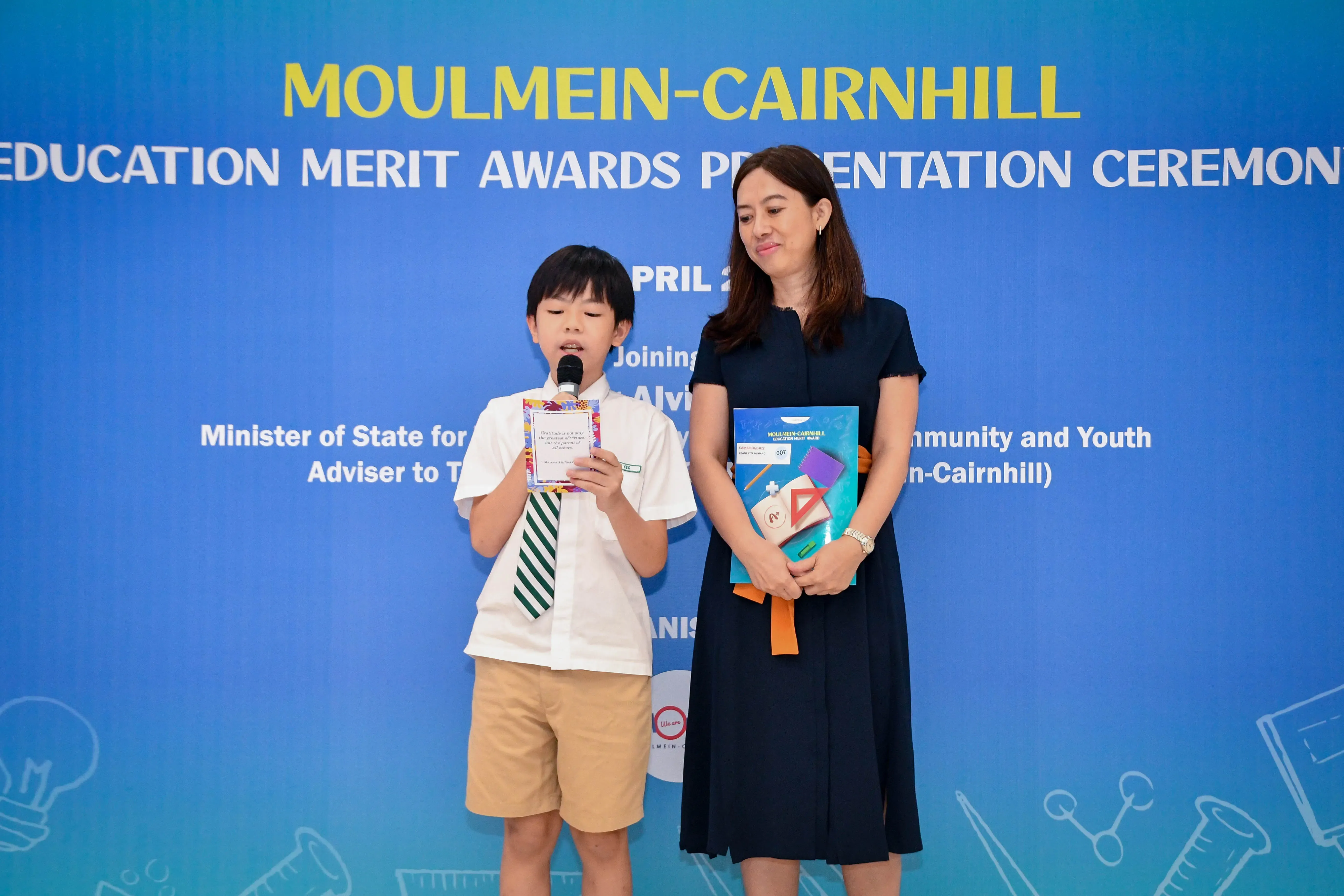 A young boy in school uniform speaks into a microphone beside a woman at the Moulmein-Cairnhill Education Merit Awards ceremony.