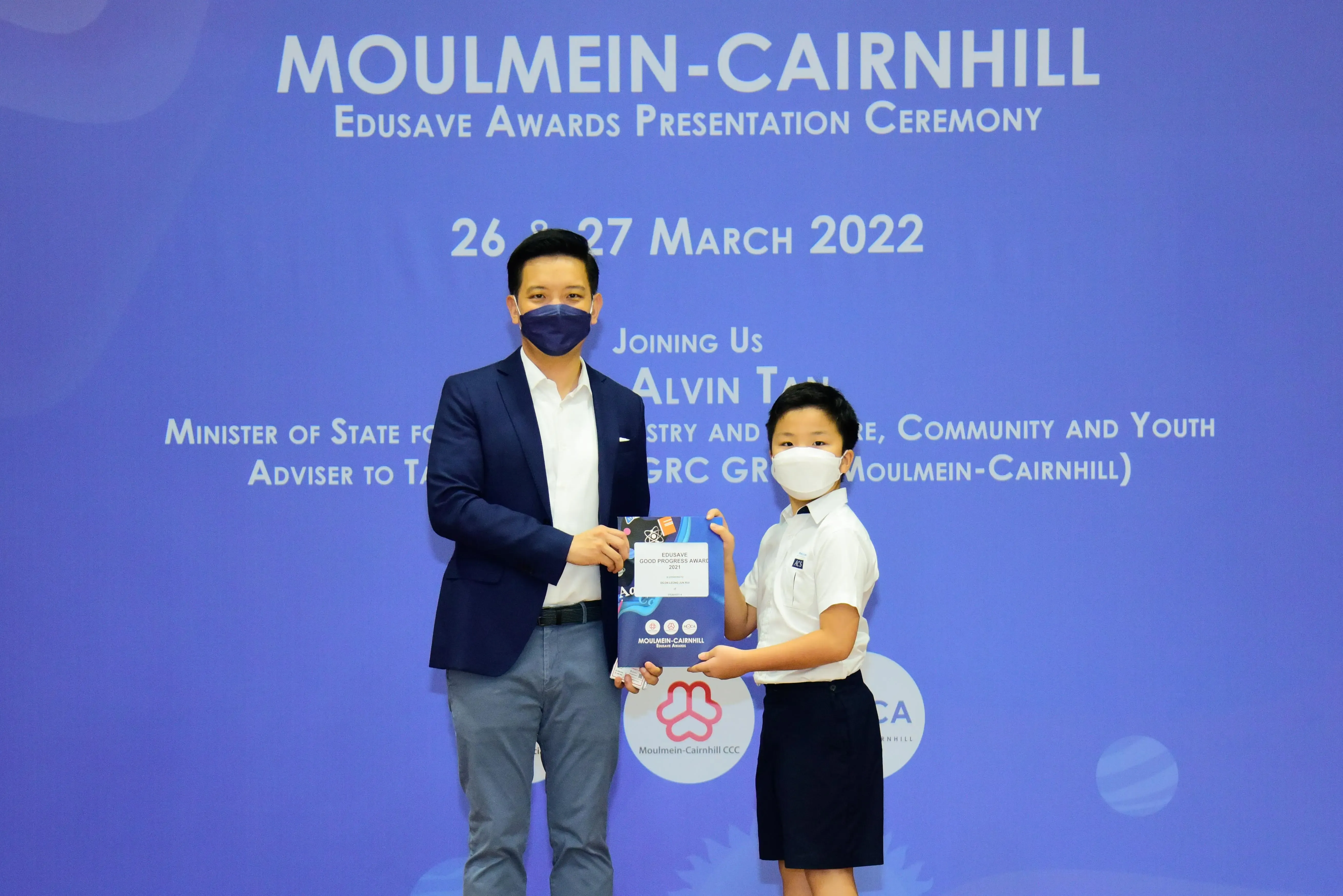 An adult and a child in masks hold an award certificate at the Moulmein-Cairnhill Edusave Awards Ceremony, March 2022.