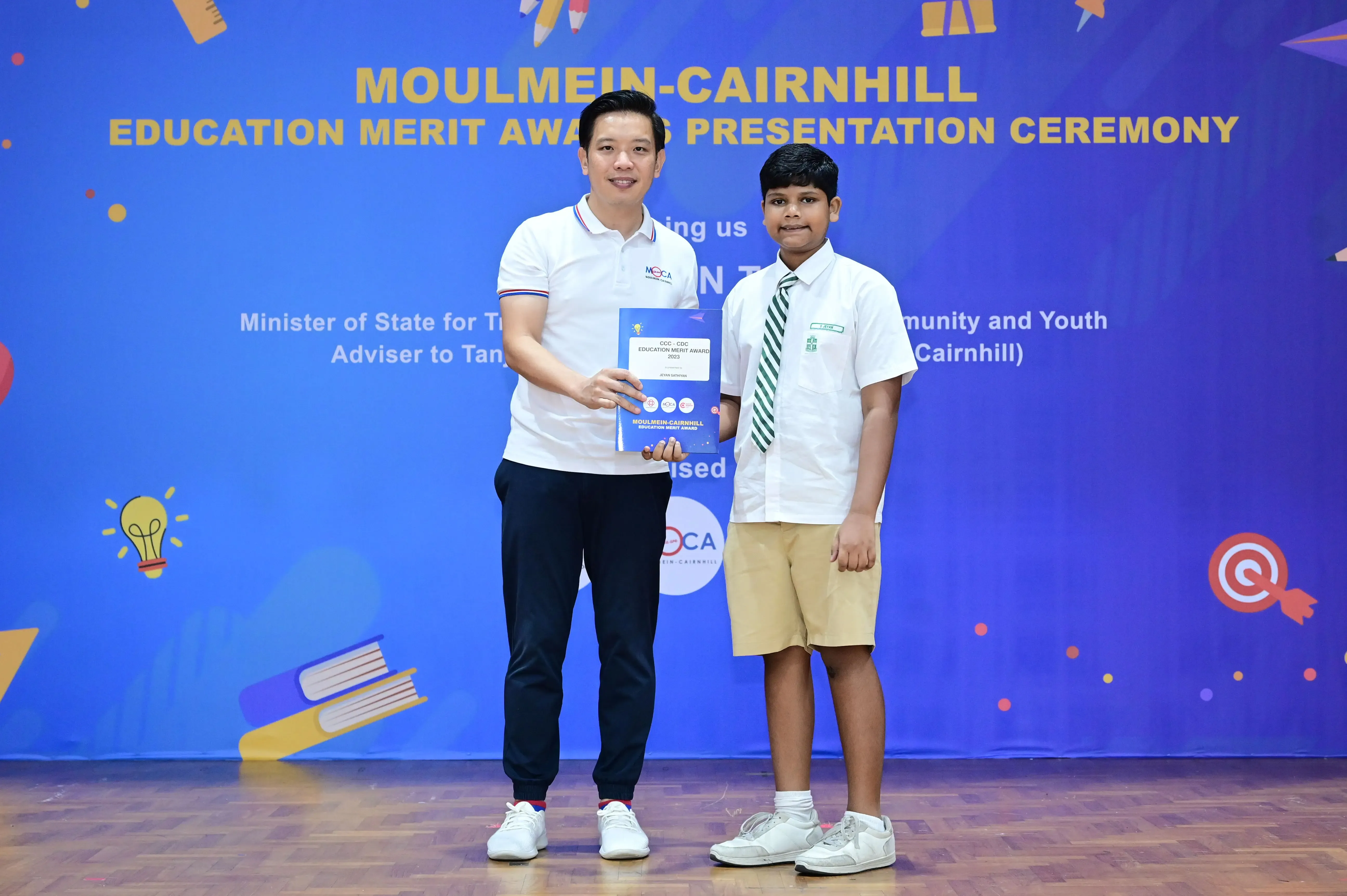 A man and a young boy in school uniform hold an award certificate at the Moulmein-Cairnhill Education Merit Award ceremony.
