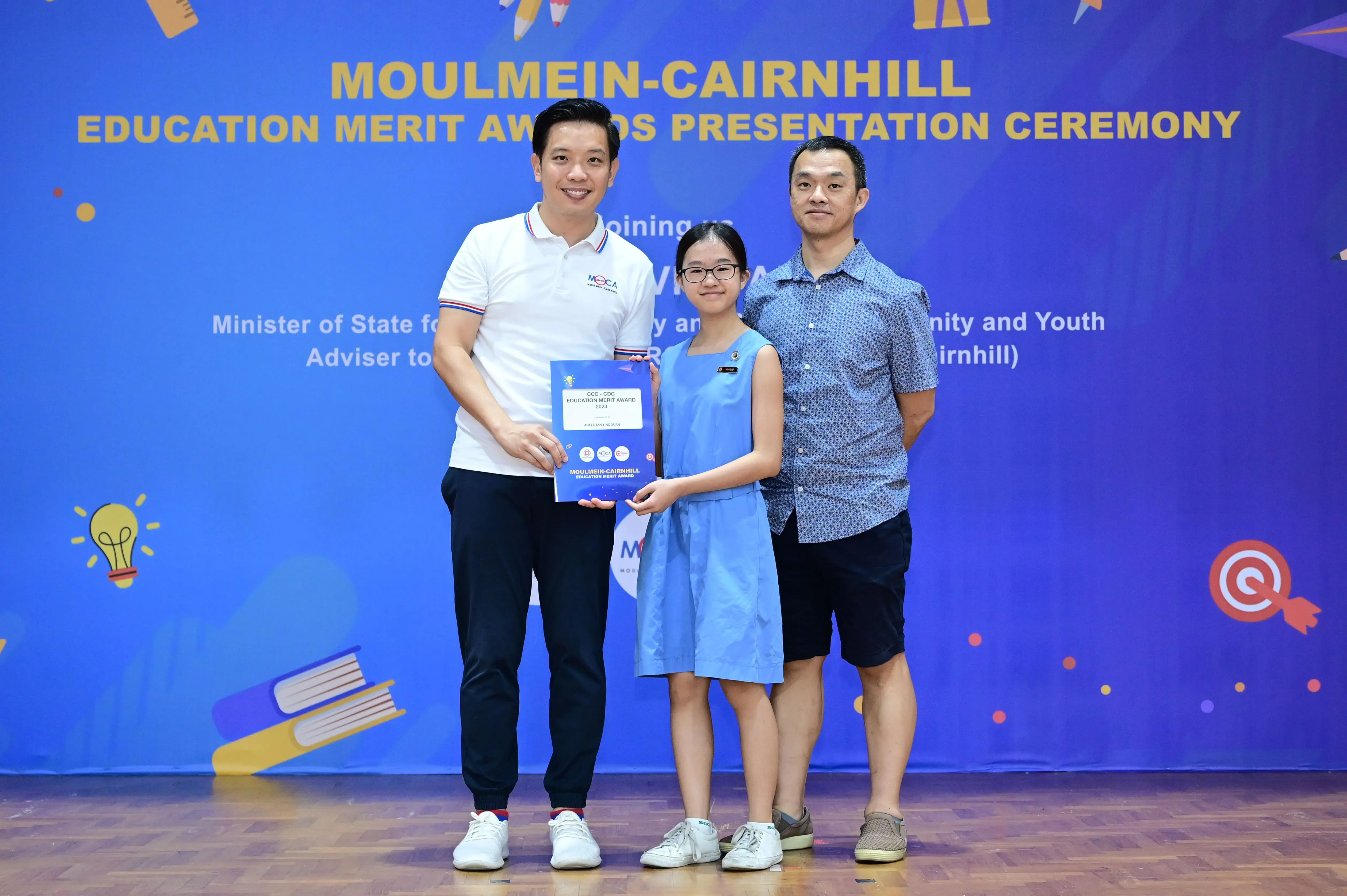 Two men and a young girl holding an award certificate at the Moulmein-Cairnhill Education Merit Awards ceremony.