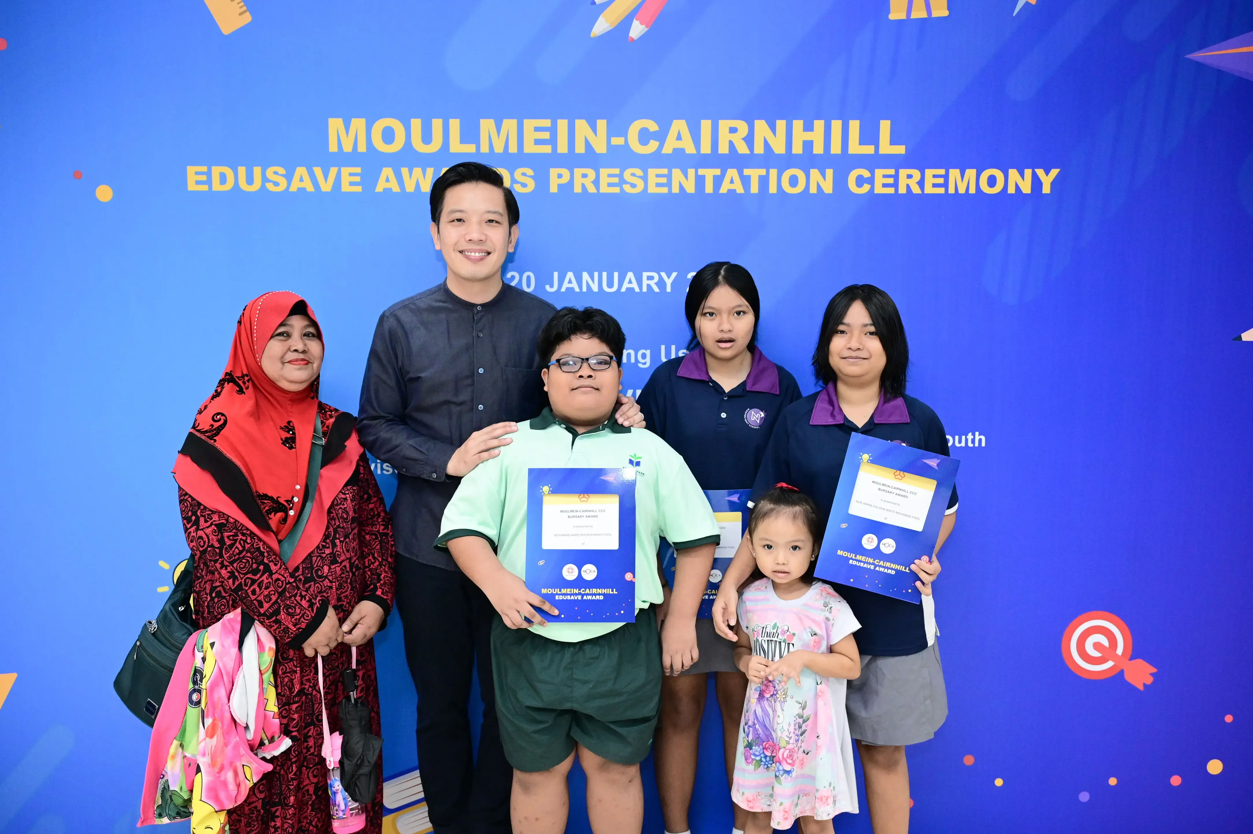 Six people pose in front of a blue Moulmein-Cairnhill Edusave Awards Presentation Ceremony banner.