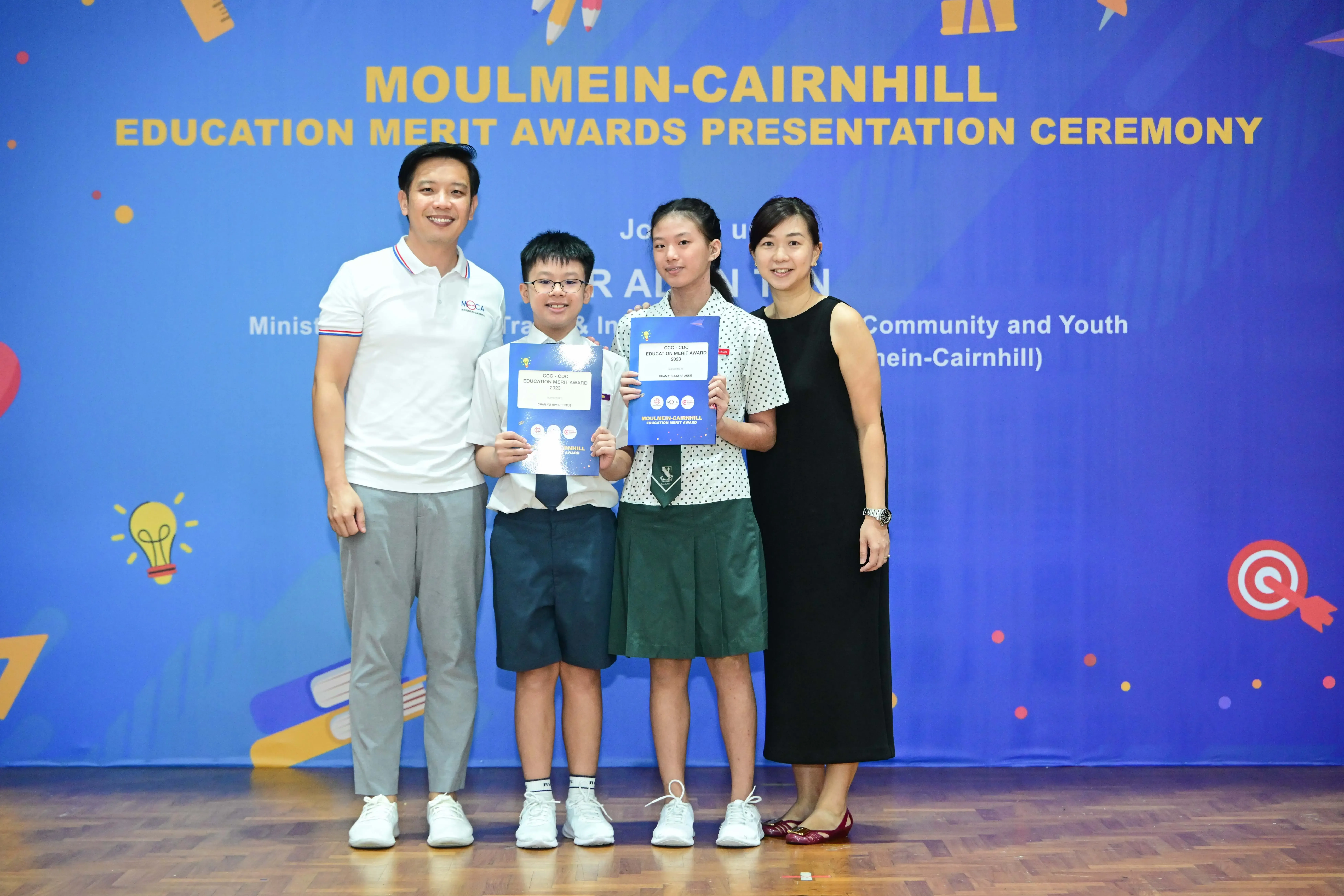 Two students holding award certificates stand with two adults at the Moulmein-Cairnhill Education Merit Awards ceremony.