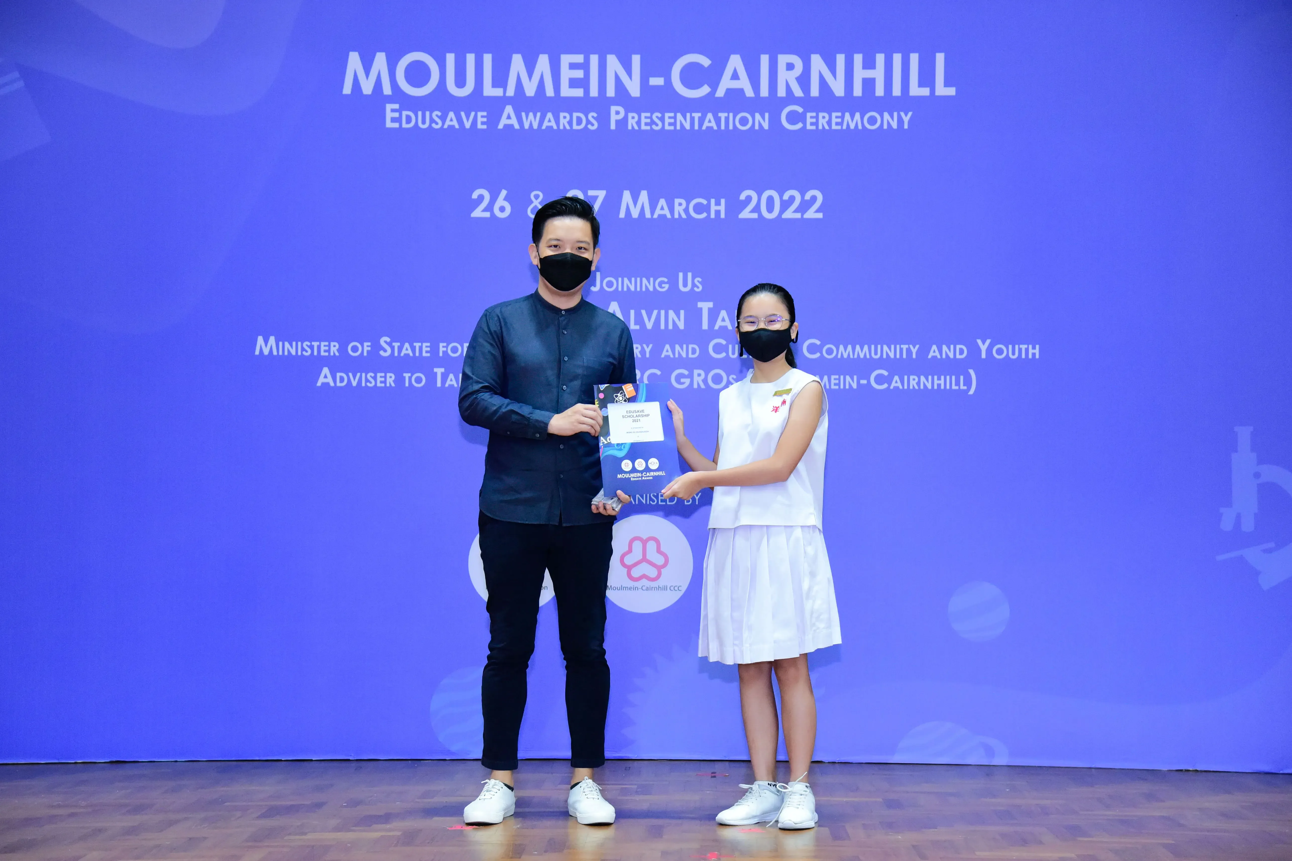 A man and a young girl in masks hold a certificate on stage at the Moulmein-Cairnhill Edusave Awards Ceremony 2022.