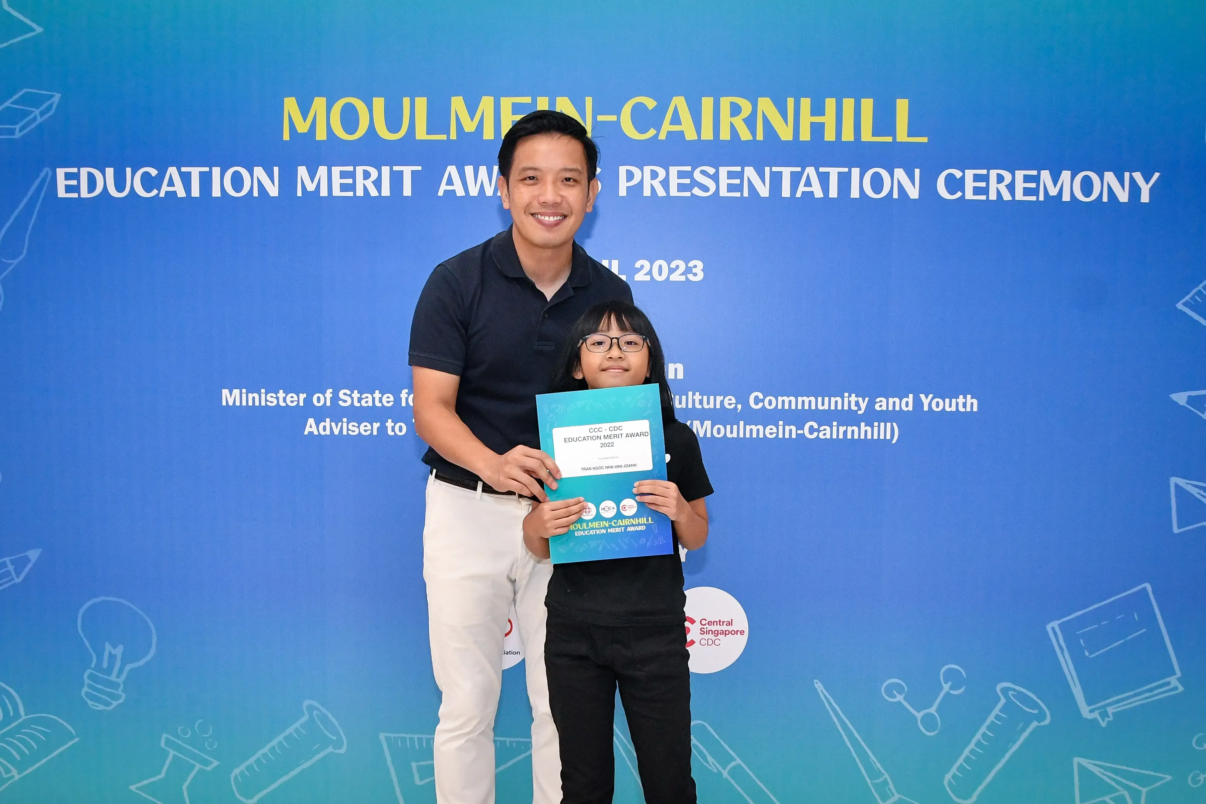 A man and a young child holding an award certificate pose at the 2023 Moulmein-Cairnhill Education Merit Award ceremony.