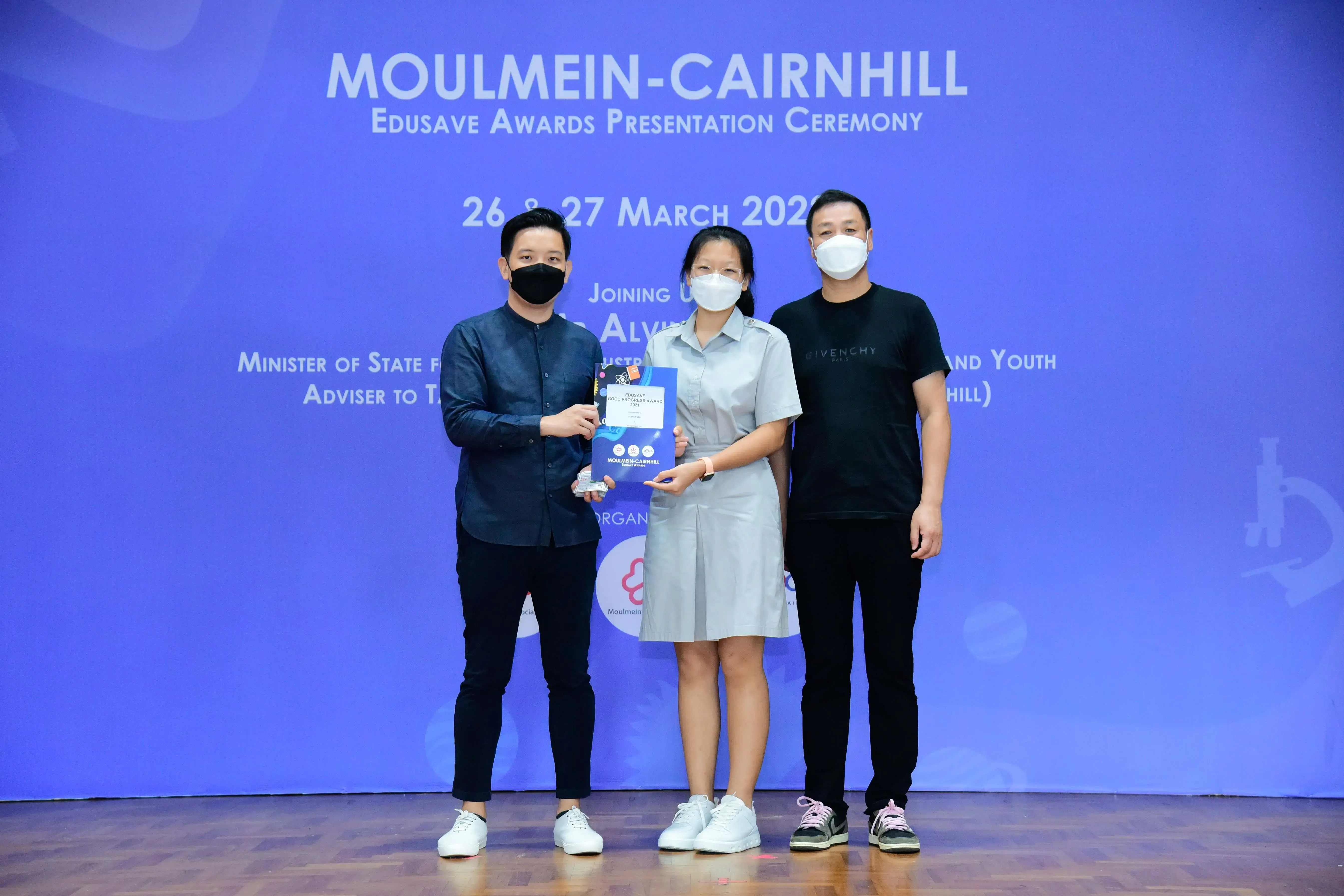 Three masked people pose on a stage at the Moulmein-Cairnhill Edusave Awards Presentation Ceremony, holding an award.