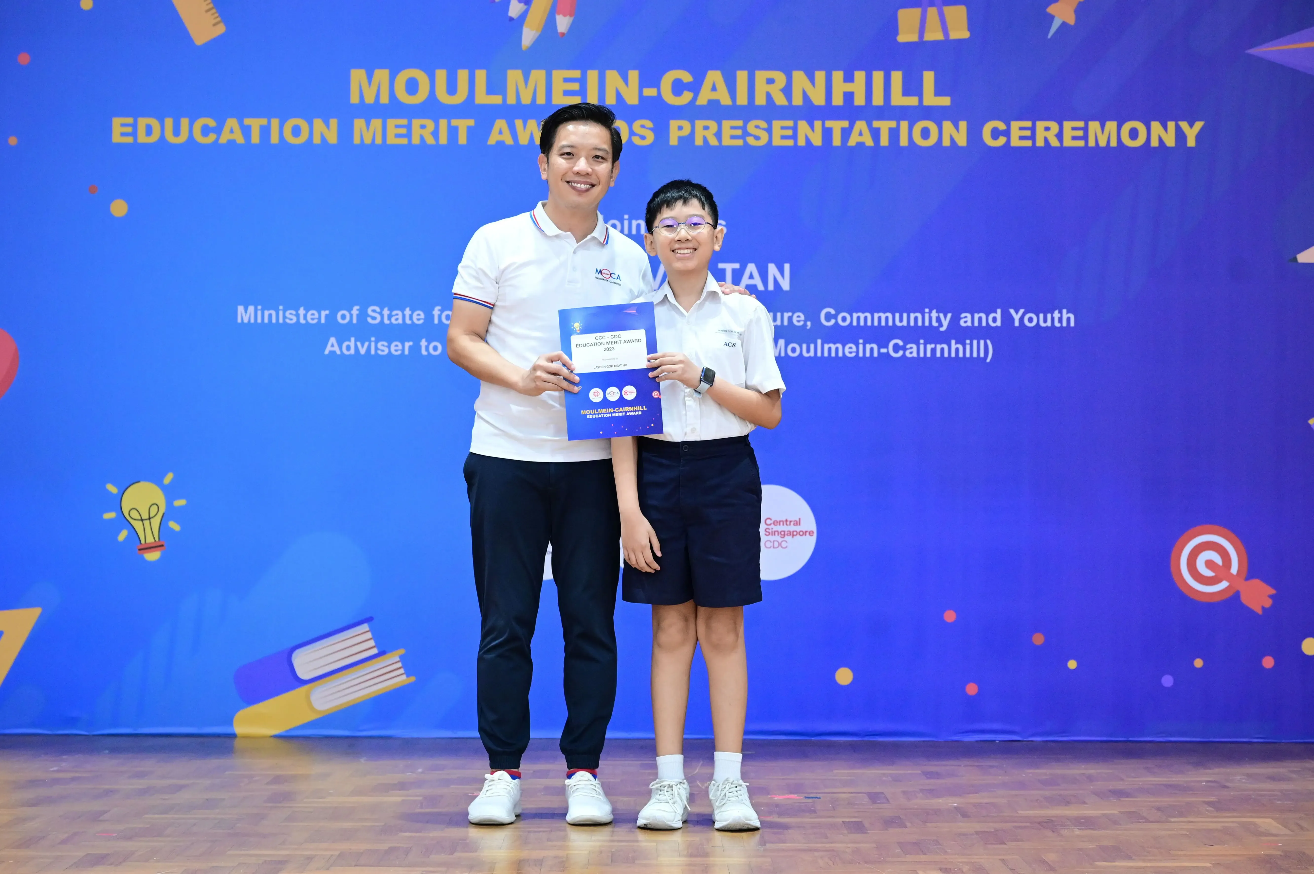 An adult and a young student hold a certificate at the Moulmein-Cairnhill Education Merit Awards Presentation Ceremony.