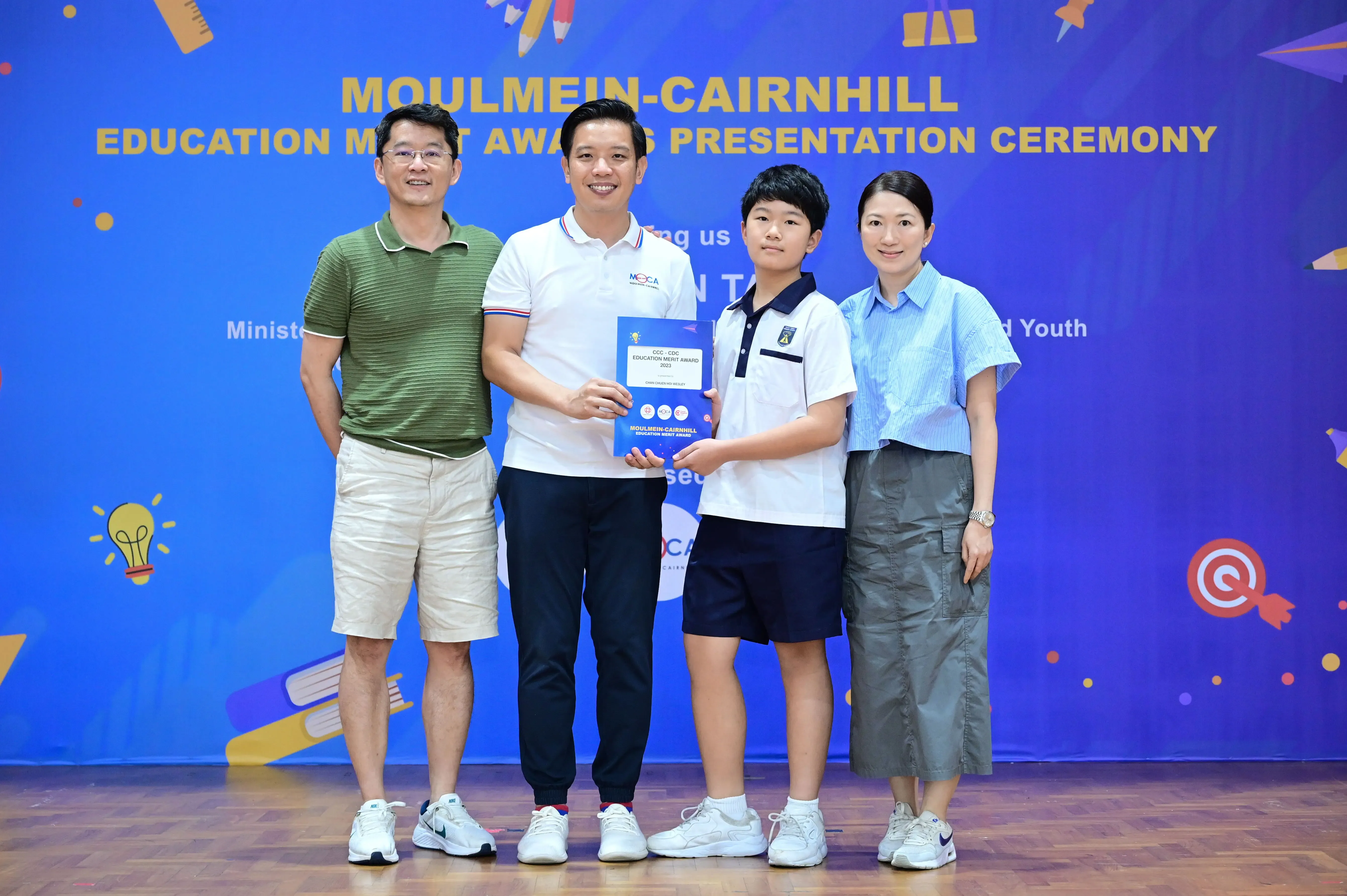Four people pose at the Moulmein-Cairnhill Education Merit Awards Presentation Ceremony, a student holds an award booklet.