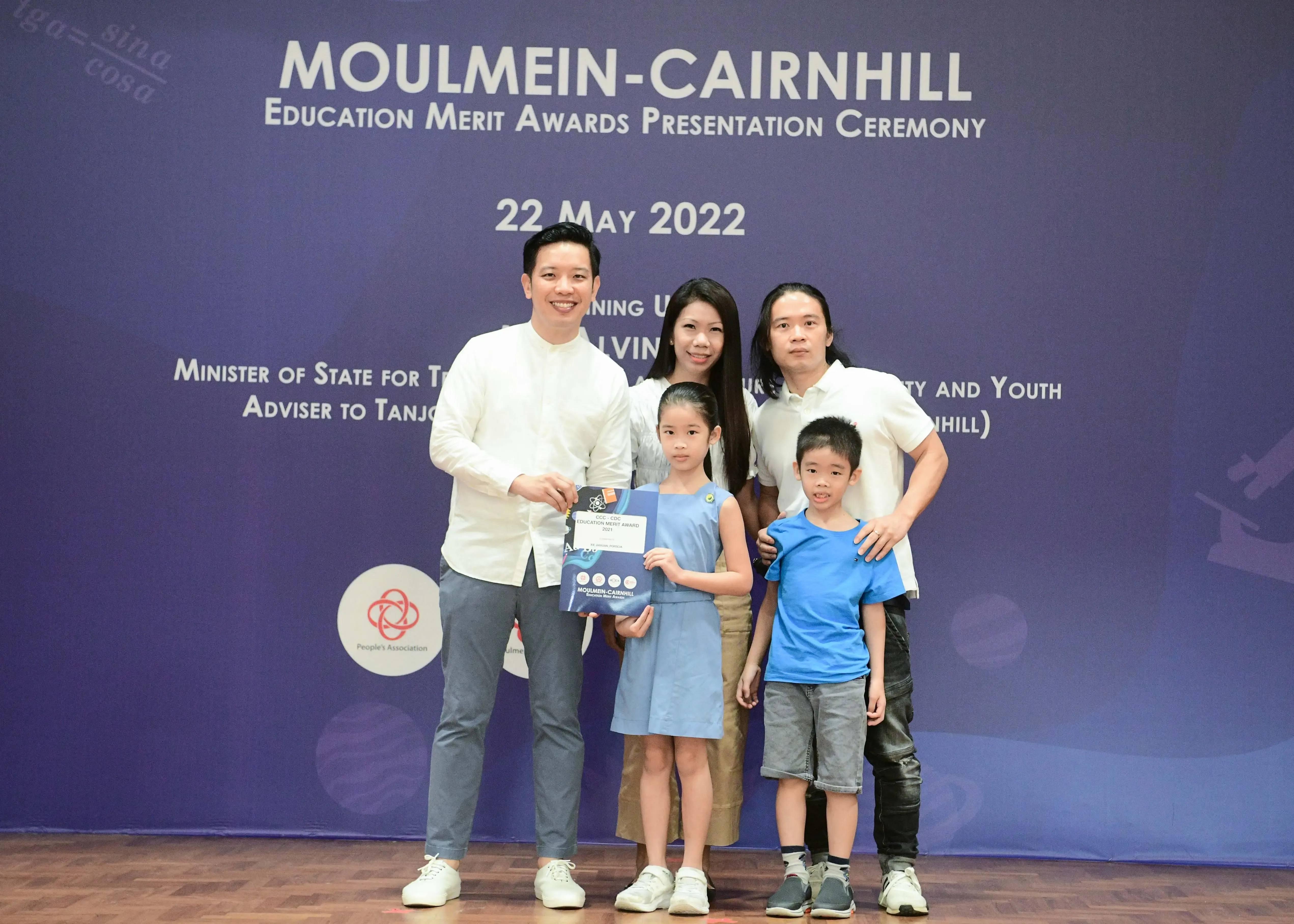 Five people pose before a Moulmein-Cairnhill Education Merit Awards banner dated 22 May 2022, with a girl holding an award.