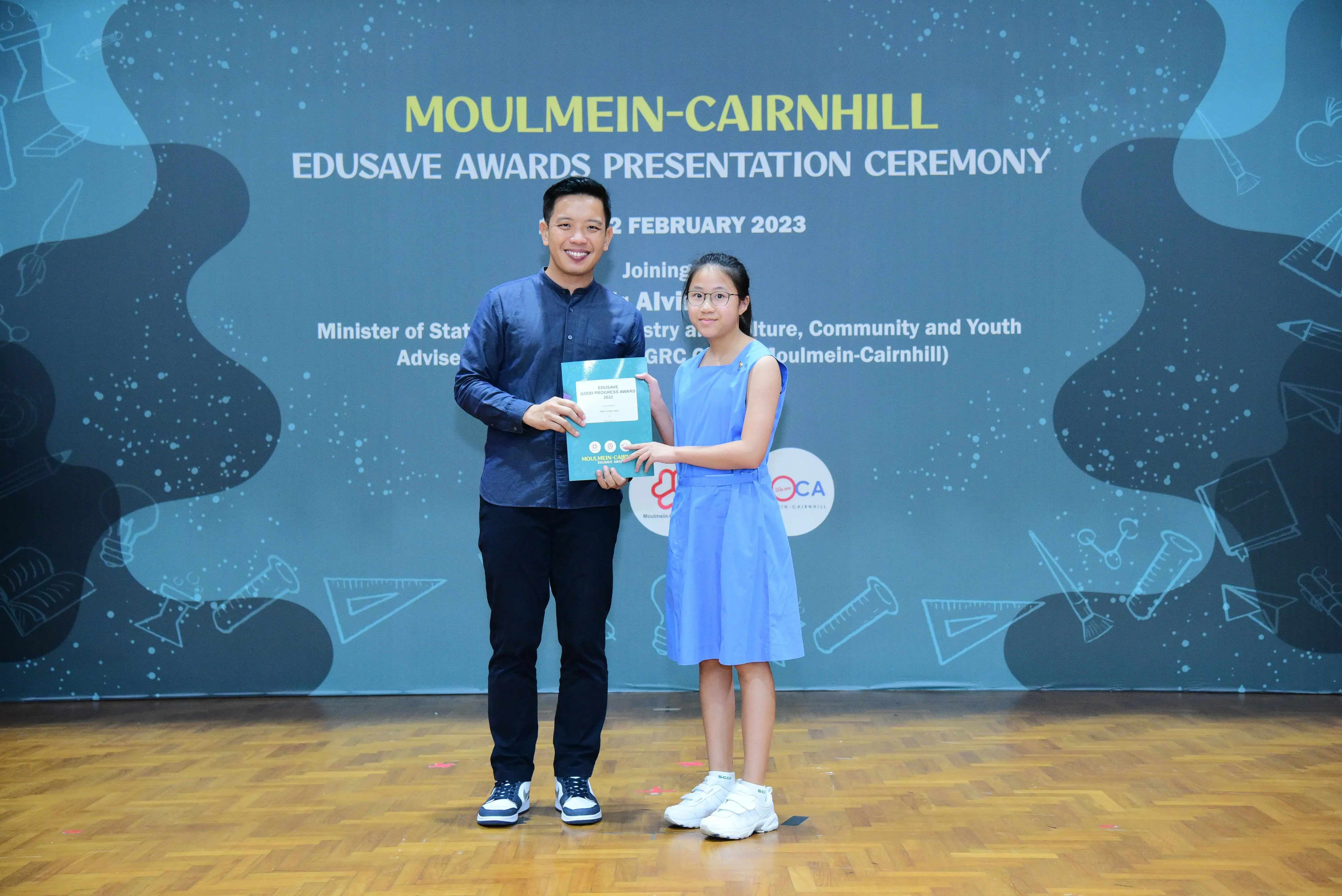 A man and a young girl hold a certificate together on stage at the Moulmein-Cairnhill Edusave Awards Ceremony 2023.
