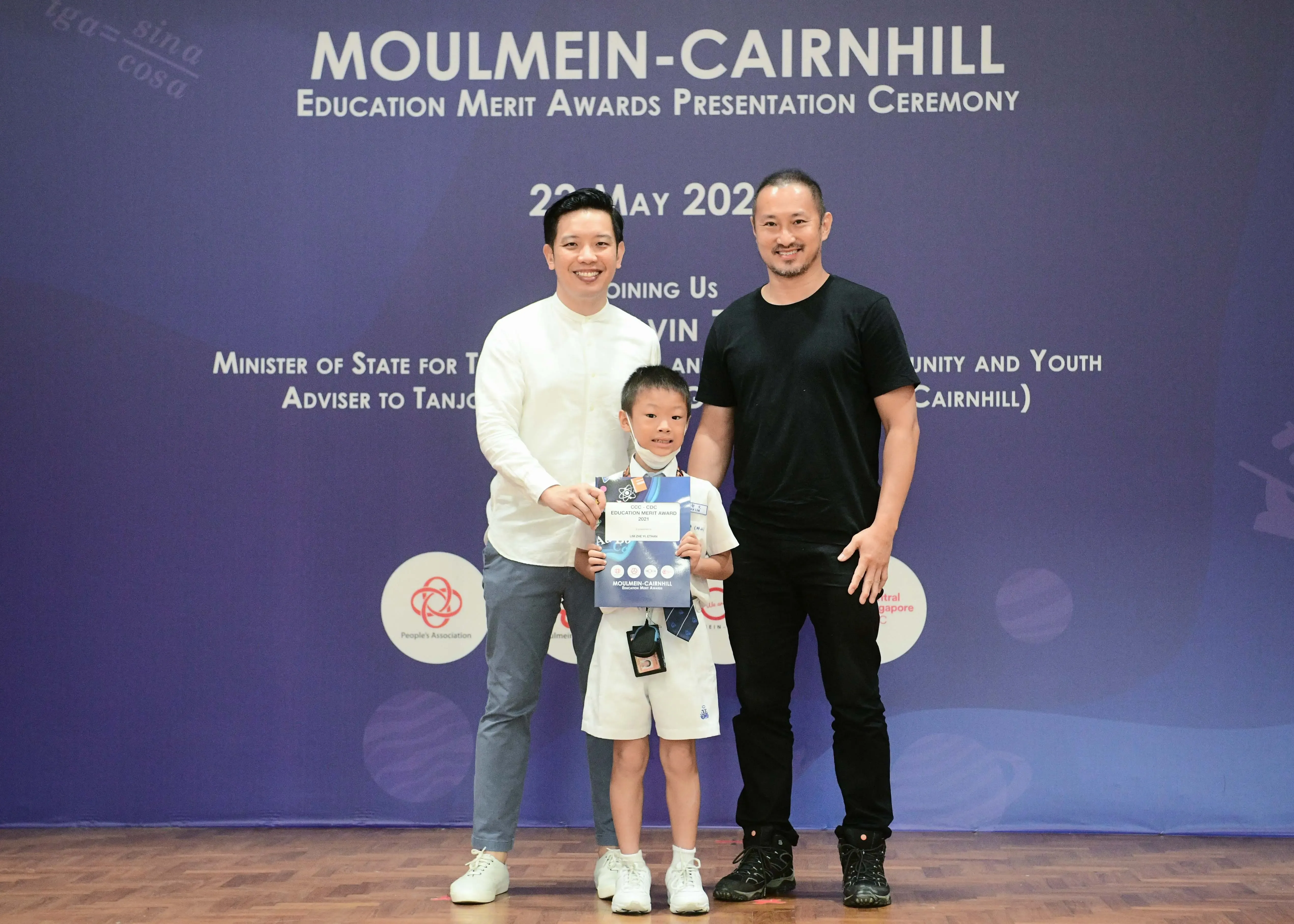 Two men and a young boy holding an award certificate pose before a Moulmein-Cairnhill Education Merit Awards banner.